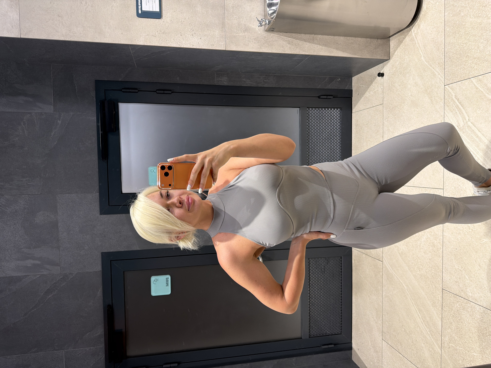 In an indoor setting with grey tiled walls and floor, a woman with blonde hair pulled back takes a selfie in a full-length mirror. She is wearing a grey, form-fitting ribbed athletic outfit consisting of a halter top and leggings. Her left hand is on her hip, and she is holding an orange smartphone in her right hand to take the photo. Behind her, two dark doors with textured metal grilles are visible, one with a light blue sign depicting a shower icon and text. To the right, a stainless steel trash receptacle with a curved top is positioned against a light-colored tiled wall.