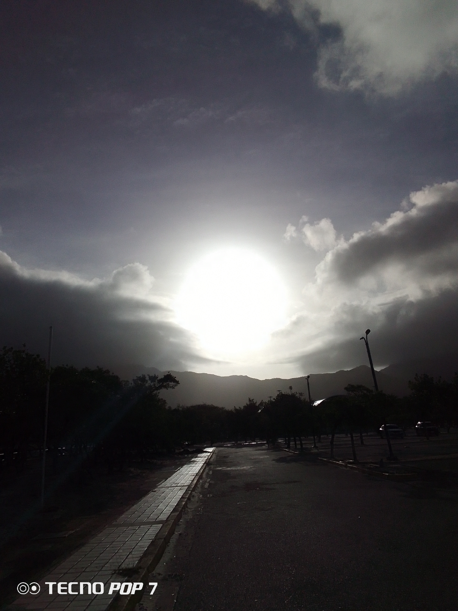 The image shows an outdoor scene in Pedro González, Venezuela, at what appears to be sunset or sunrise. The sky is predominantly covered with clouds, and the sun is a bright, circular orb shining through a gap in the clouds. The scene has a high contrast, with dark shadows in the foreground and around the edges of the frame. A paved path runs along the left side of the frame, and an asphalt road extends into the distance. Trees and other vegetation line the sides of the road. In the background, mountains are silhouetted against the bright sunlight. Several street lights are visible. There are two vehicles further down the path. The words "Tecno POP 7" are printed at the bottom.