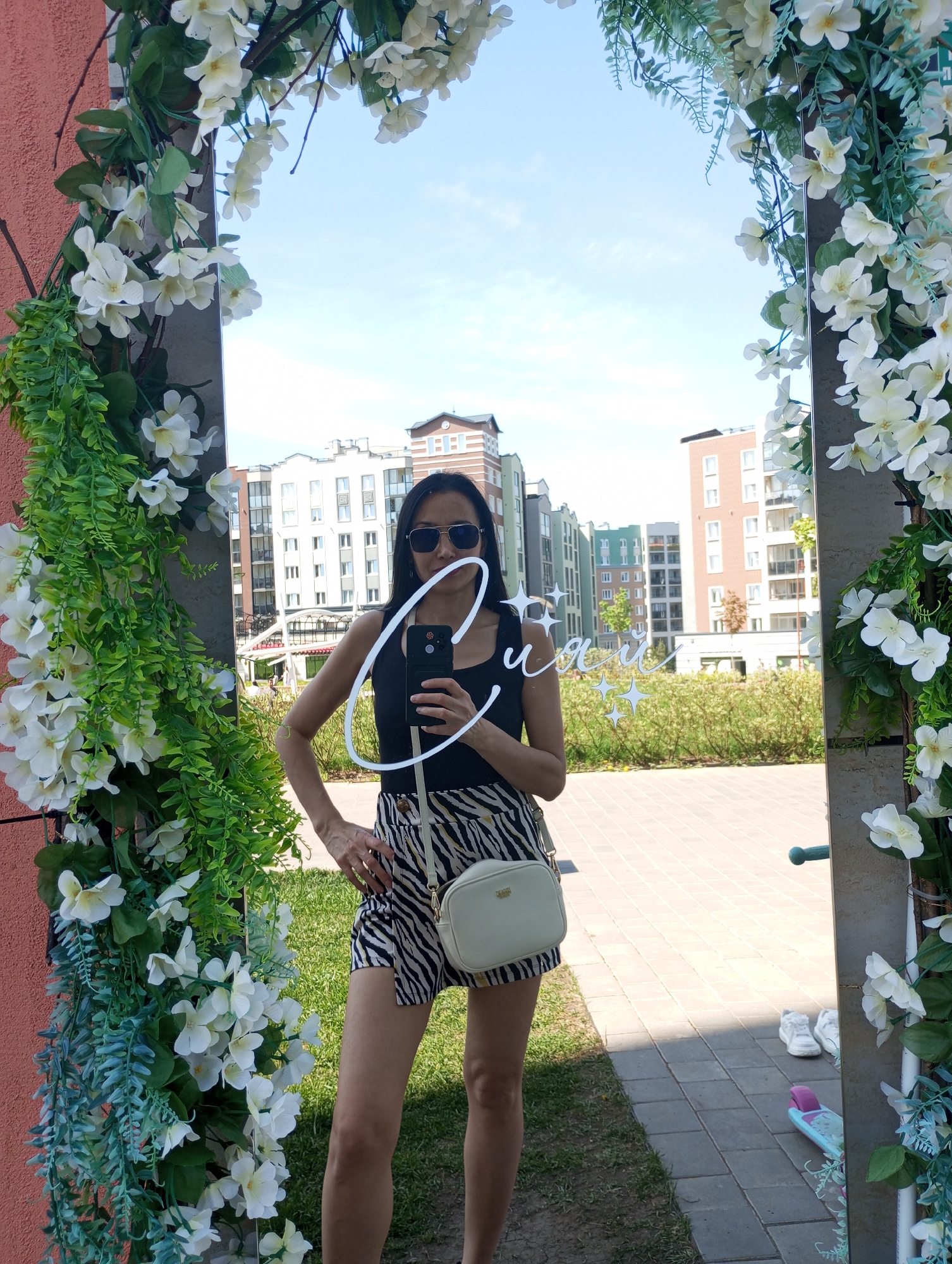 A woman is standing in front of a mirror taking a selfie. The mirror is decorated with white flowers and greenery. The woman is wearing a black tank top, zebra-striped shorts, sunglasses, and a white purse. She is holding a black smartphone with a white light ring attached. In the background, there are apartment buildings, green grass, and trees. The pavement beneath her is made of grey stone. In the lower right of the mirror's reflection, a pair of white shoes and a scooter are visible. The location is Izhevsk, Russia.