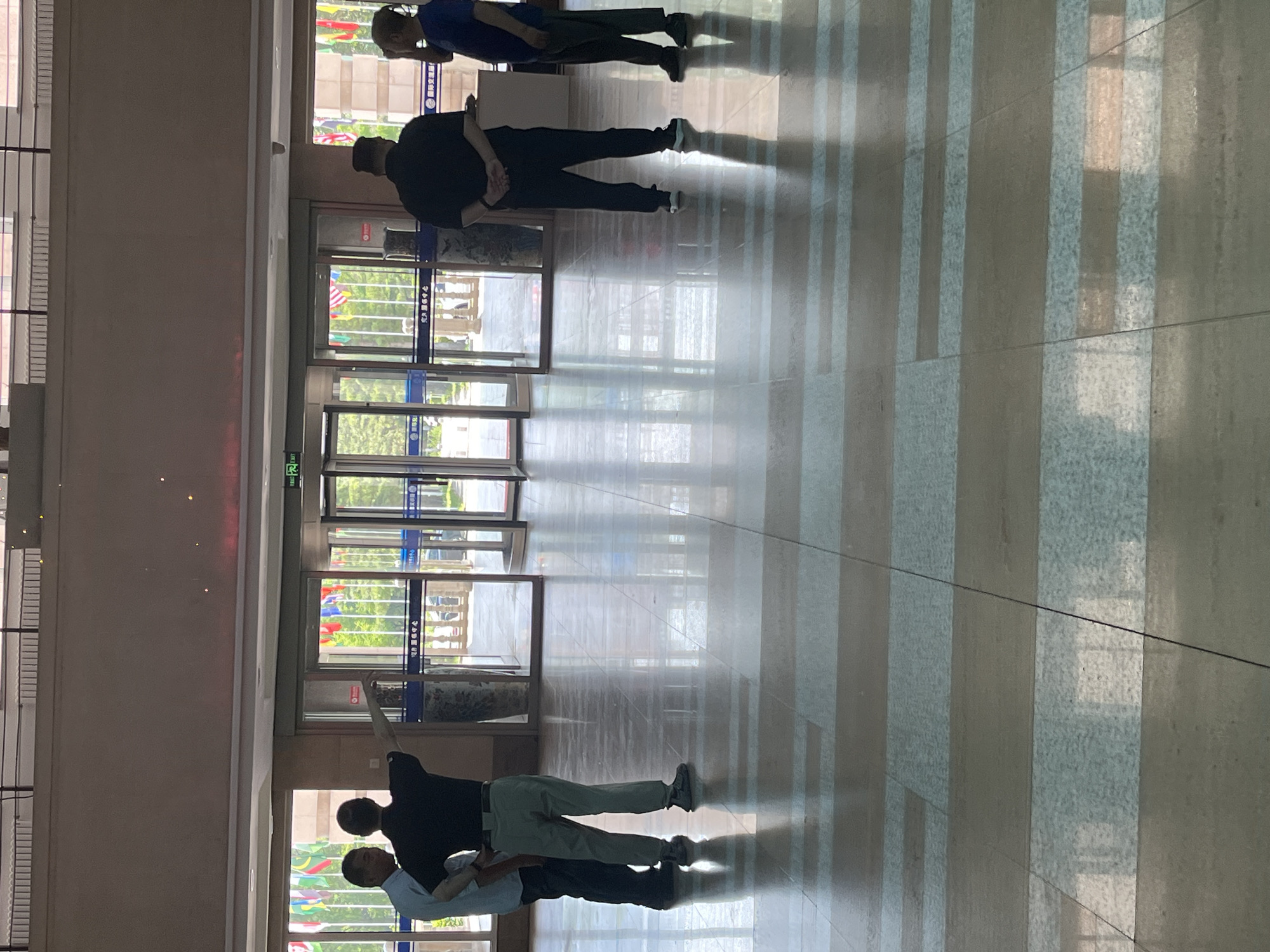 The image shows an indoor scene, possibly a lobby or entrance hall, with a polished tiled floor. There are several people present, and a set of glass double doors leading outside, where flags are visible.
In the foreground, the floor is made of large rectangular tiles in alternating bands of beige and light blue/grey. The floor reflects the light and the objects in the room.
There are four men visible. On the left, two men are standing and talking, with one gesturing towards the doors. The man on the left is wearing a light blue shirt and dark pants, and the man on the right is wearing a dark t-shirt and light-colored pants.
Further into the background, towards the right of the image, two more men are standing. One is wearing a black t-shirt and black pants, with his hands clasped behind his back. The other man is wearing a dark blue t-shirt and dark pants.
Behind the men, there are large glass double doors, some of which appear to be automatic revolving doors. Through the doors, an outdoor area is visible with trees and numerous national flags displayed on poles. The flags suggest a location that may represent international relations or an event. Above the doors, a green sign with white text, likely indicating an exit, is visible.
The overall environment appears to be a well-lit, modern public building. The architectural style and the presence of multiple national flags could indicate a government building, an international organization's office, or a venue hosting an international event in Changchun City, China.