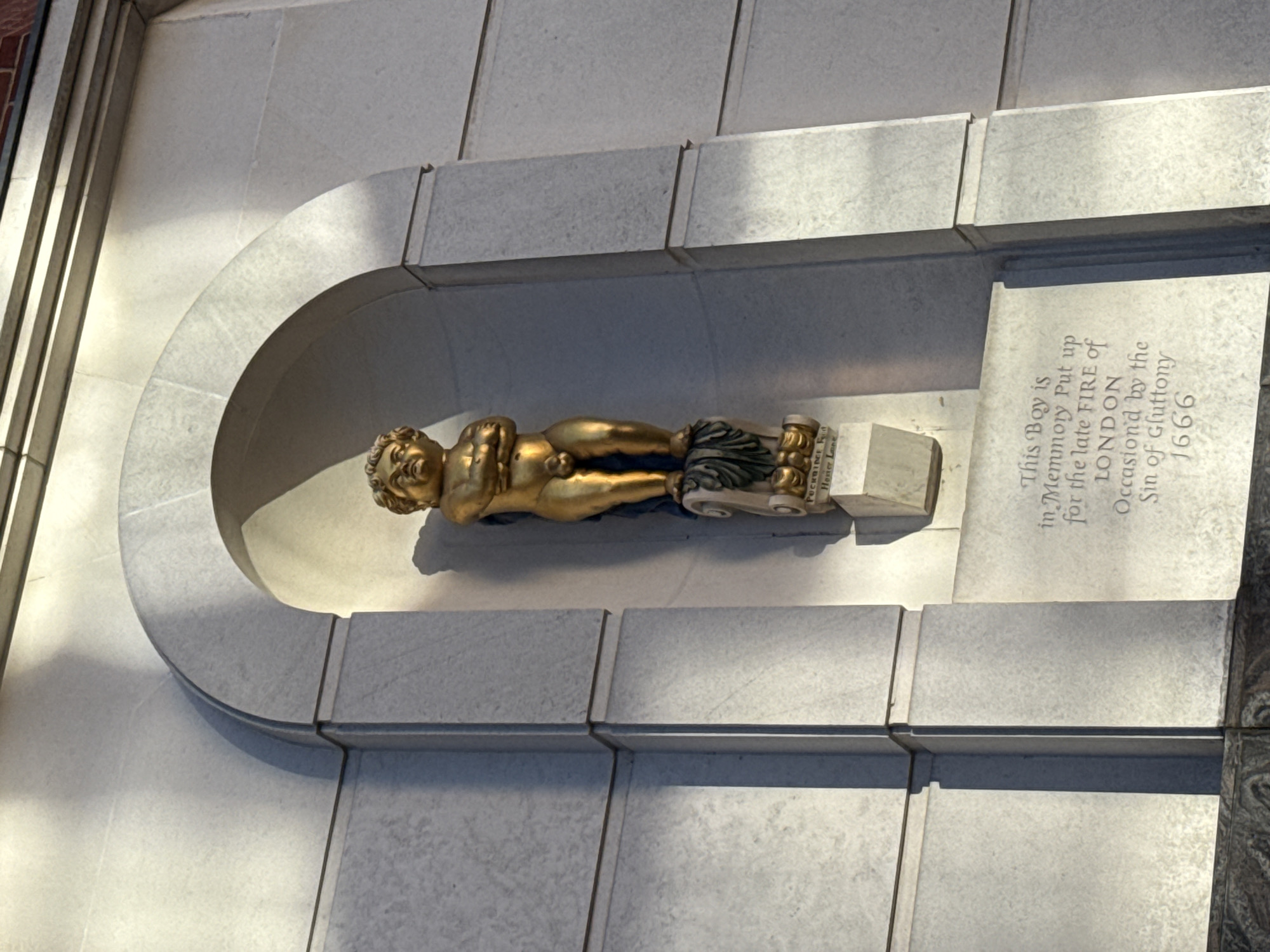 The image shows a gilded statue of a naked boy lying on his side within a niche in a light gray stone wall. The statue is supported on a stone plinth. Next to the niche, an inscription is visible on a stone plaque, which reads: "This Boy is in Memory Put up for the late FIRE of LONDON Occasion'd by the Sin of Gluttony 1666". The location is in the city of London, United Kingdom.