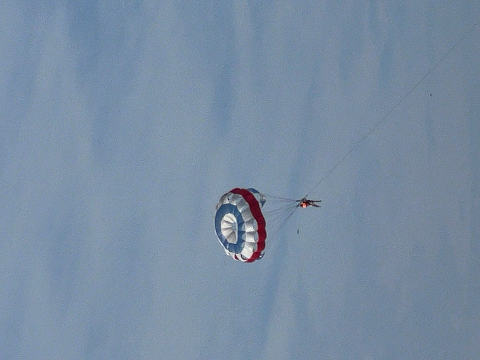 A person is parasailing in the sky above the city of Đà Nẵng, Vietnam. The parasail is red, white, and blue. The person is wearing an orange life vest. The sky is clear and blue.