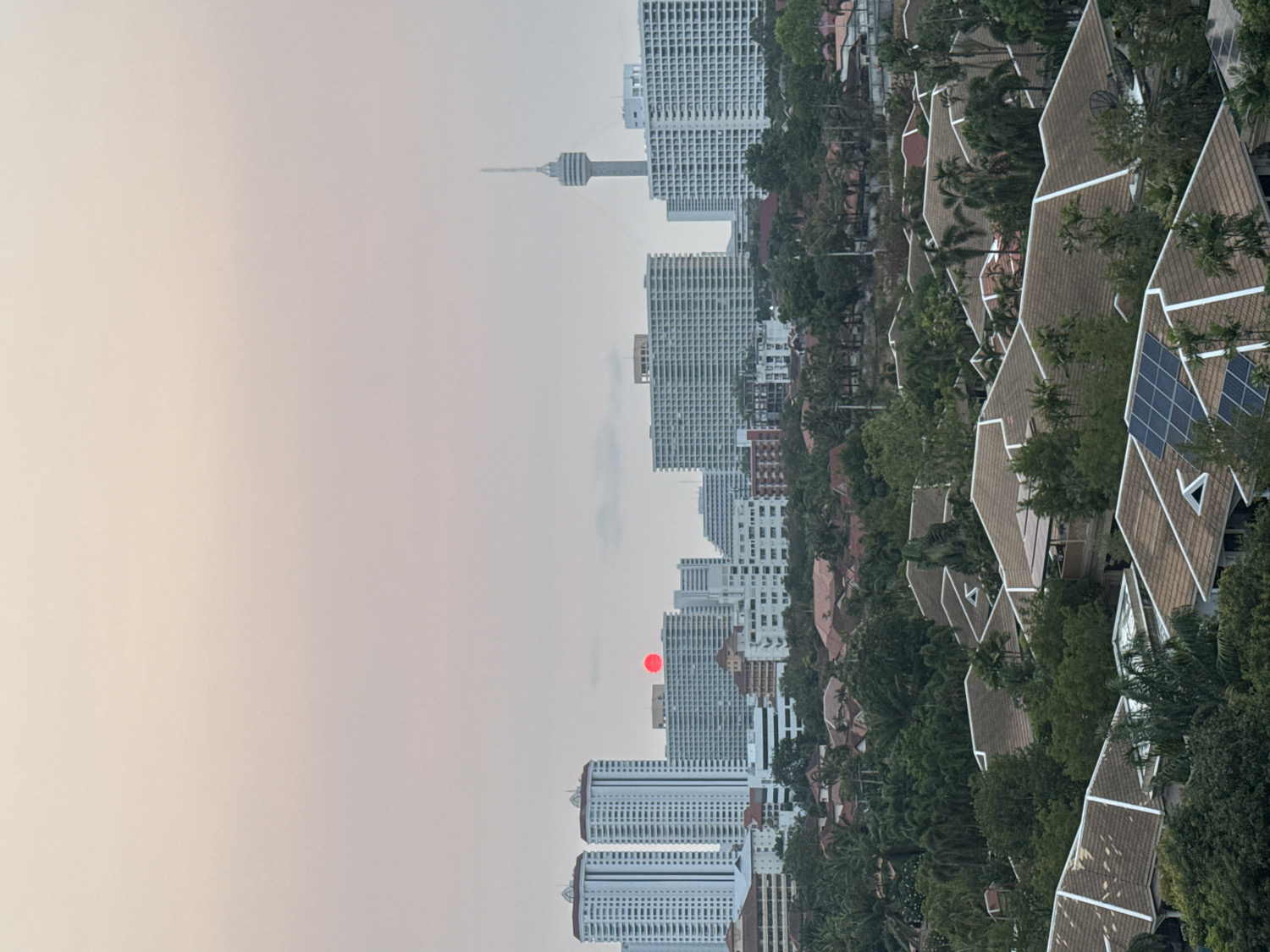 The image captures an elevated view of Pattaya City, Thailand, at either sunrise or sunset. A distinct, small, red sun is visible low in the hazy sky, positioned between several urban structures.
The scene features a diverse architectural landscape. In the foreground, on the right side of the frame, are numerous low-rise buildings with traditional Thai-style, steeply pitched and multi-tiered brown roofs. These structures are nestled amidst abundant lush green foliage and palm trees, with several roofs clearly displaying blue solar panels.
Behind these traditional buildings, the city transitions into a dense concentration of modern high-rise buildings, characterized by their rectangular forms and numerous windows. These multi-story structures extend into the background, creating a layered urban panorama. A prominent landmark, the Pattaya Park Tower, a slender observation tower, stands tall in the mid-ground, silhouetted against the atmospheric sky.
The overall weather appears hazy or overcast, contributing to the soft, diffused light across the scene. No specific human activity is visible, and there is no discernible text in the image. The dominant colors include the muted grays and whites of the buildings, the rich greens of the tropical vegetation, and the warm, soft tones of the hazy sky, accented by the striking red sun.