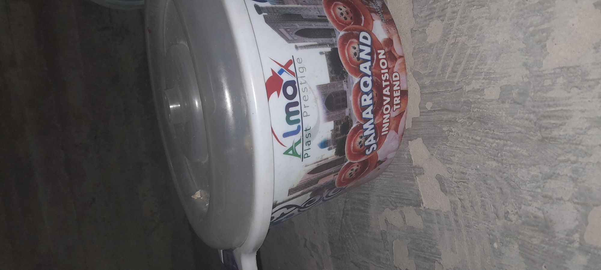 The image shows a white plastic container with a clear lid, featuring a graphic design. The design includes the logo "Almax Plast Prestige" and an image of what appears to be a historical building complex, likely in Uzbekistan, given the text "SAMARQAND INNOVATSION TREND" printed on the container. The container is placed on a rough, textured surface that appears to be a wall or ground with chipping paint or plaster. The lighting is dim, casting shadows and suggesting an indoor or dimly lit outdoor environment. There are no people or animals visible in the image, nor any specific indication of the city of Chartak.