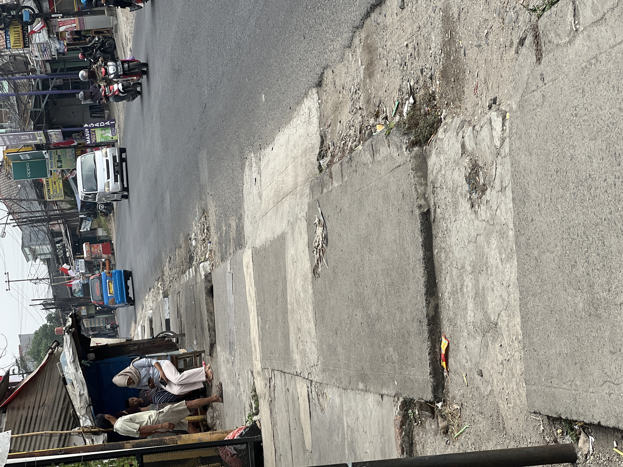 The image captures a street scene in Citeureup, Indonesia. A man and a child are standing next to a small roadside structure, possibly a stall or shop, with a woman sitting on a stool in front of it. The woman is wearing a hijab and is looking down at her phone. Further down the street, a blue van is parked on the side of the road, and several motorcycles are passing by. The road itself shows signs of wear and tear, with some patches of broken asphalt and debris. The environment appears to be an urban or semi-urban area with a mix of small businesses and residential buildings visible in the background. The sky is overcast, suggesting a possibly humid or rainy climate.
