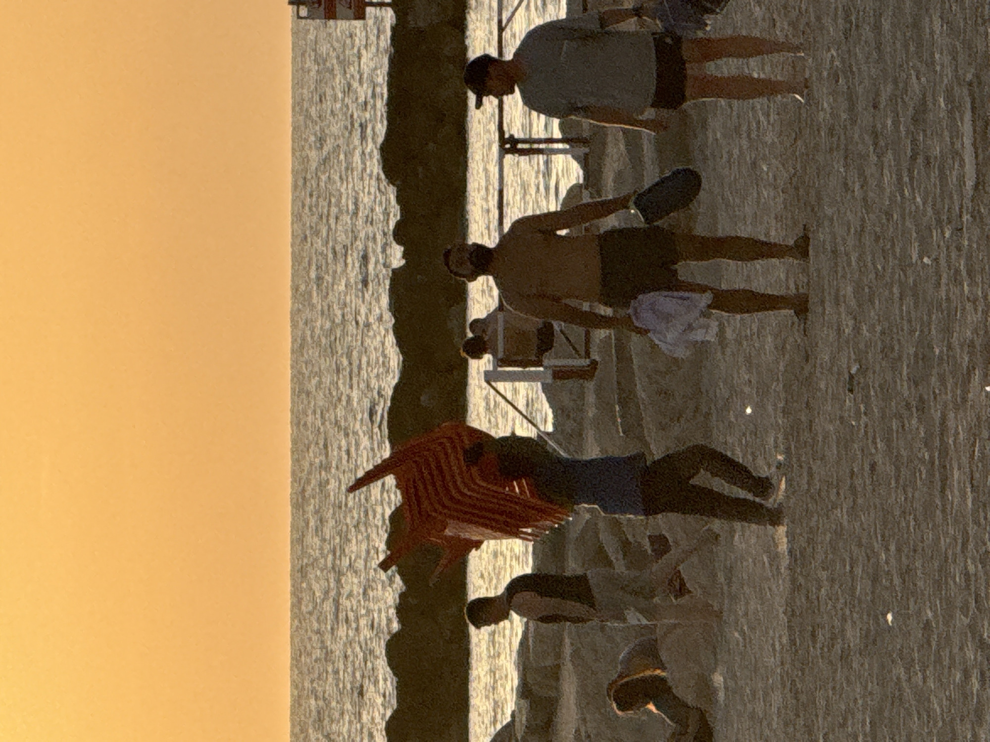 The image depicts a beach scene in Tel-Aviv, Israel, during sunset. The sky is a warm orange hue, and the sea is visible in the background. Several people are present on the sandy beach. In the foreground, two men are walking. One is shirtless, wearing dark shorts, and carrying a dark object in his right hand and a white garment in his left. The other man is wearing a light-colored t-shirt and dark shorts, also with a dark object in his hand. To their left, a person wearing a blue shirt is carrying a stack of red chairs on their head. Further to the left, a man in a dark vest and light pants is standing. In the middle ground, there are several rocks and a railing. A person is seated on a lounge chair in the distance. A person with long hair is lying on the sand in the left portion of the image. The overall atmosphere suggests a relaxed end-of-day gathering at the beach.