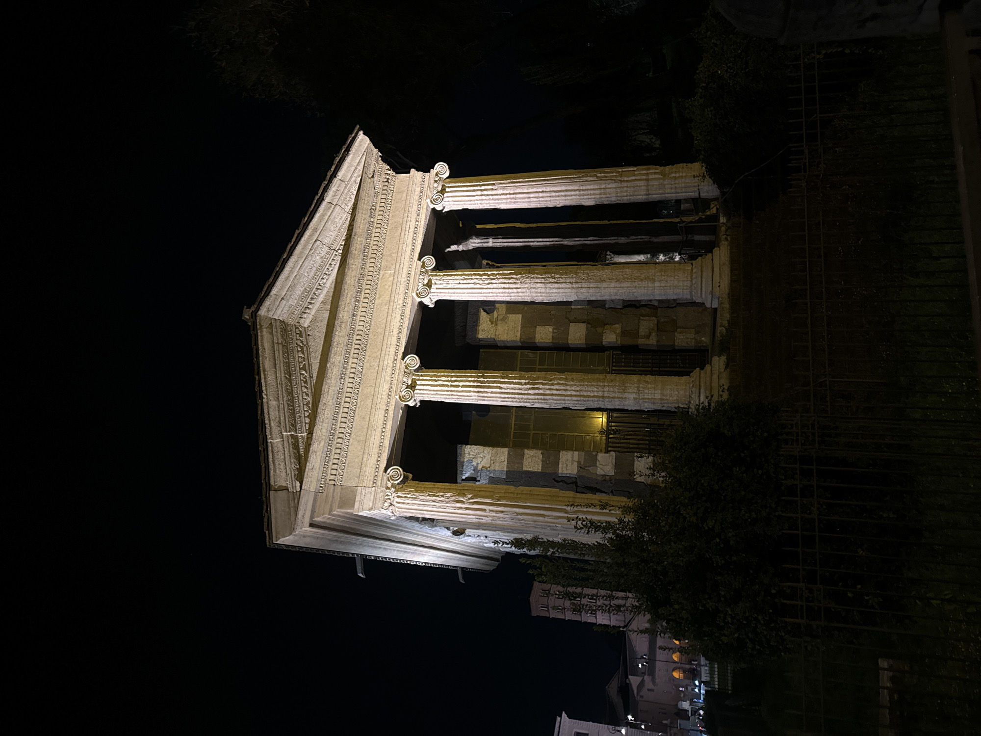 The image shows the Temple of Portunus in Rome, Italy, illuminated at night. The temple is a well-preserved Roman temple dedicated to the god Portunus. It is a rectangular structure with a portico supported by Ionic columns. The building is illuminated with bright lights. The temple is located in the Forum Boarium, an area near the Tiber River. Trees and a fence are on the right side of the image. Buildings can be seen in the background on the bottom right.