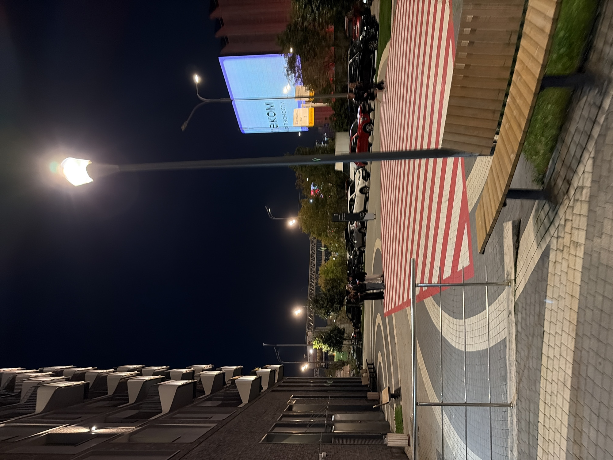 The image depicts a pedestrian crossing at night, likely in Moscow, Russia, given the Cyrillic text on a large LED billboard in the background. The billboard displays the word "ТЕКОМ" and other Cyrillic characters, suggesting a commercial or informational context. The pedestrian crossing is marked with alternating red and white stripes.
To the left, a modern building with a brick facade and balconies is visible, illuminated by streetlights. Several streetlights are also present in the scene, casting light on the pedestrian area and the parked cars. A prominent lamppost stands in the foreground, partially obscuring the view of the billboard.
A group of three individuals, two women and one person of indeterminate gender, are standing near the pedestrian crossing, appearing to be in conversation. Another individual is seen walking across the red and white stripes in the distance.
Several cars are parked in a lot behind the pedestrian crossing, including a white sedan, a red SUV, and black sedans. Wooden benches are situated near the building and along the walkway. The ground is paved with interlocking bricks, and a metal railing runs along the edge of the walkway. The overall atmosphere is that of a public urban space at night, with elements of modern architecture and commercial advertising.