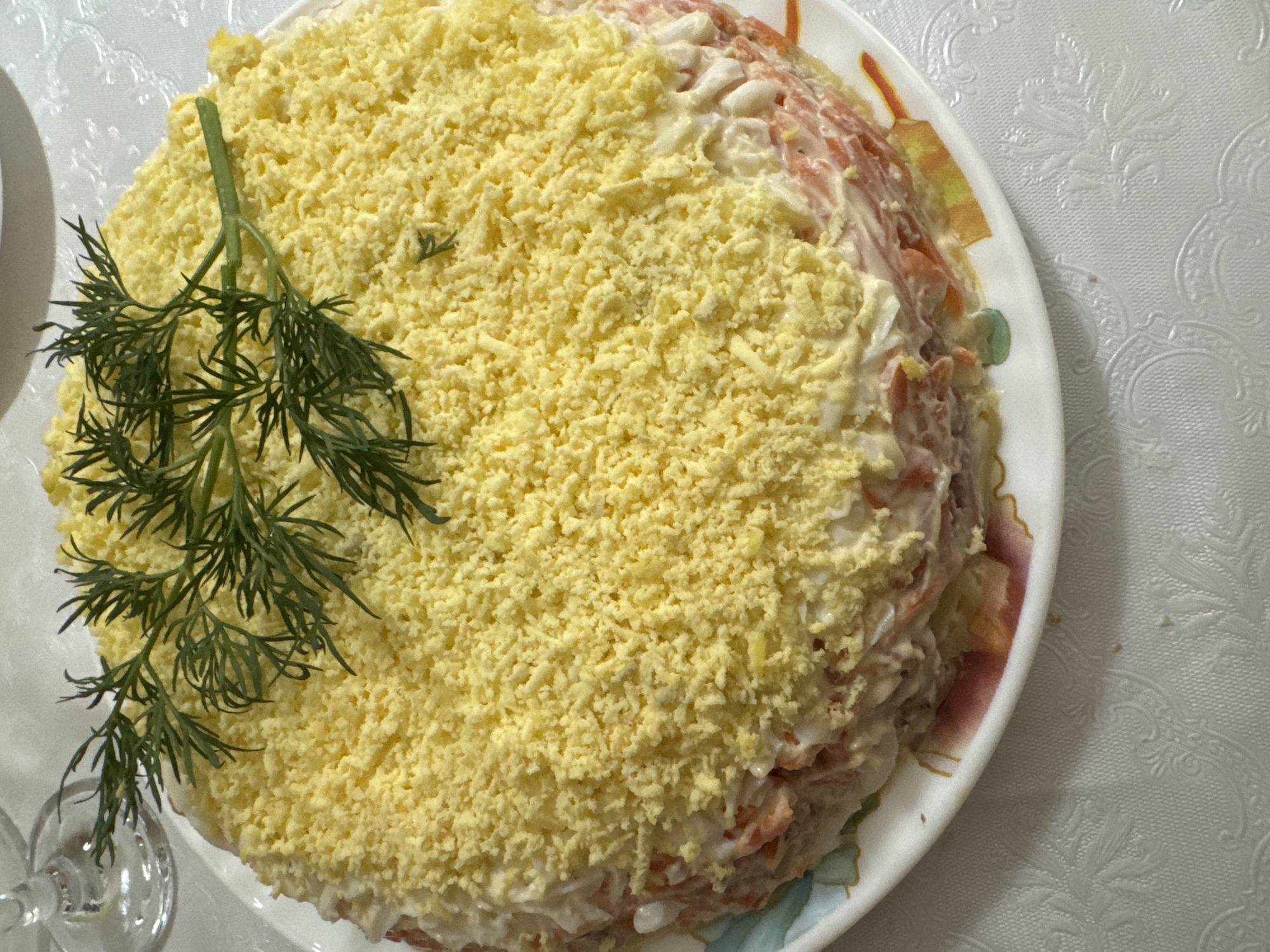 A top-down shot displays a multilayered salad garnished with dill on a decorative plate. The salad's top layer is predominantly yellow, likely shredded egg yolk. Visible below are layers with ingredients possibly including mayonnaise, carrots, and other components. The plate features a floral and geometric design on its rim. The background shows a textured white tablecloth and the blurry stem of a glass in the lower left corner. It is highly likely that this image was taken in a private setting, but without additional information, it is difficult to verify the location in Chaikovsky, Russia.