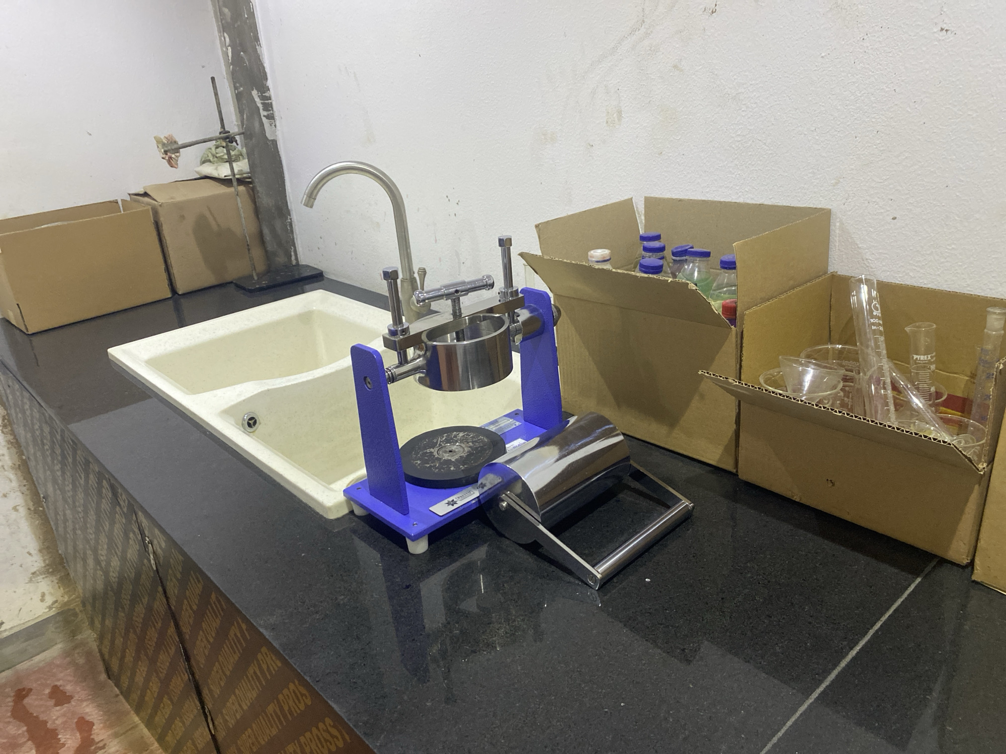 The image depicts a laboratory setting in Paipe, Nigeria, featuring a countertop sink flanked by equipment and storage boxes. On the counter, a blue and stainless steel testing apparatus stands near the sink, with a cylindrical chamber above a circular base. To the right of the apparatus are open cardboard boxes filled with laboratory glassware and bottles. Additional cardboard boxes are to the left of the sink, and a metal stand supports a piece of equipment against the wall. The countertop is dark-colored, and the wall behind the sink is light-colored and appears textured. Text is visible on the side of the countertop.