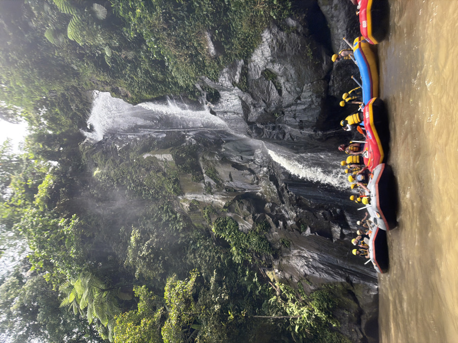 A waterfall cascades down a rocky cliff covered in lush greenery. Several colorful rafts filled with people wearing yellow helmets and life vests are positioned in a muddy river beside the waterfall. The rafts are connected in a line. The scene captures an outdoor adventure activity, likely white water rafting. The lighting appears bright, with hints of sunlight filtering through the foliage.