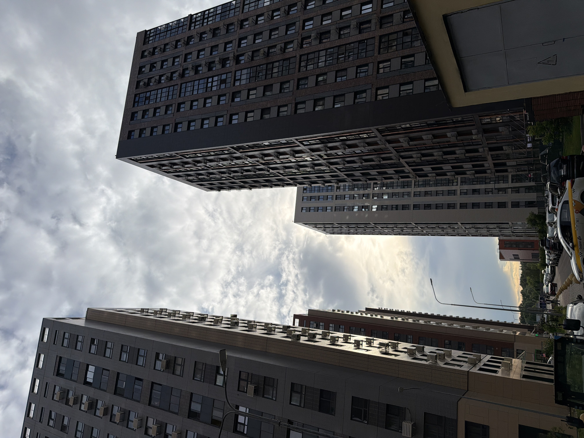 The image depicts a street view in Moscow, Russia, dominated by tall residential buildings. The architecture consists of modern, high-rise apartment complexes with numerous windows and balconies. One building on the left is clad in dark grey brick, while the building on the right features a darker facade with visible brickwork. Between these, a lighter grey building is visible. The sky above is overcast with dramatic clouds, and sunlight is breaking through in the distance, creating a bright, hazy effect. In the foreground and along the street, several cars are parked and driving, including what appear to be yellow taxis. The street is lined with trees and greenery, suggesting a residential urban environment. There is a small yellow building with a large grey garage door in the lower right of the frame. Streetlights are also visible along the sidewalk. The overall impression is that of a densely populated urban area.