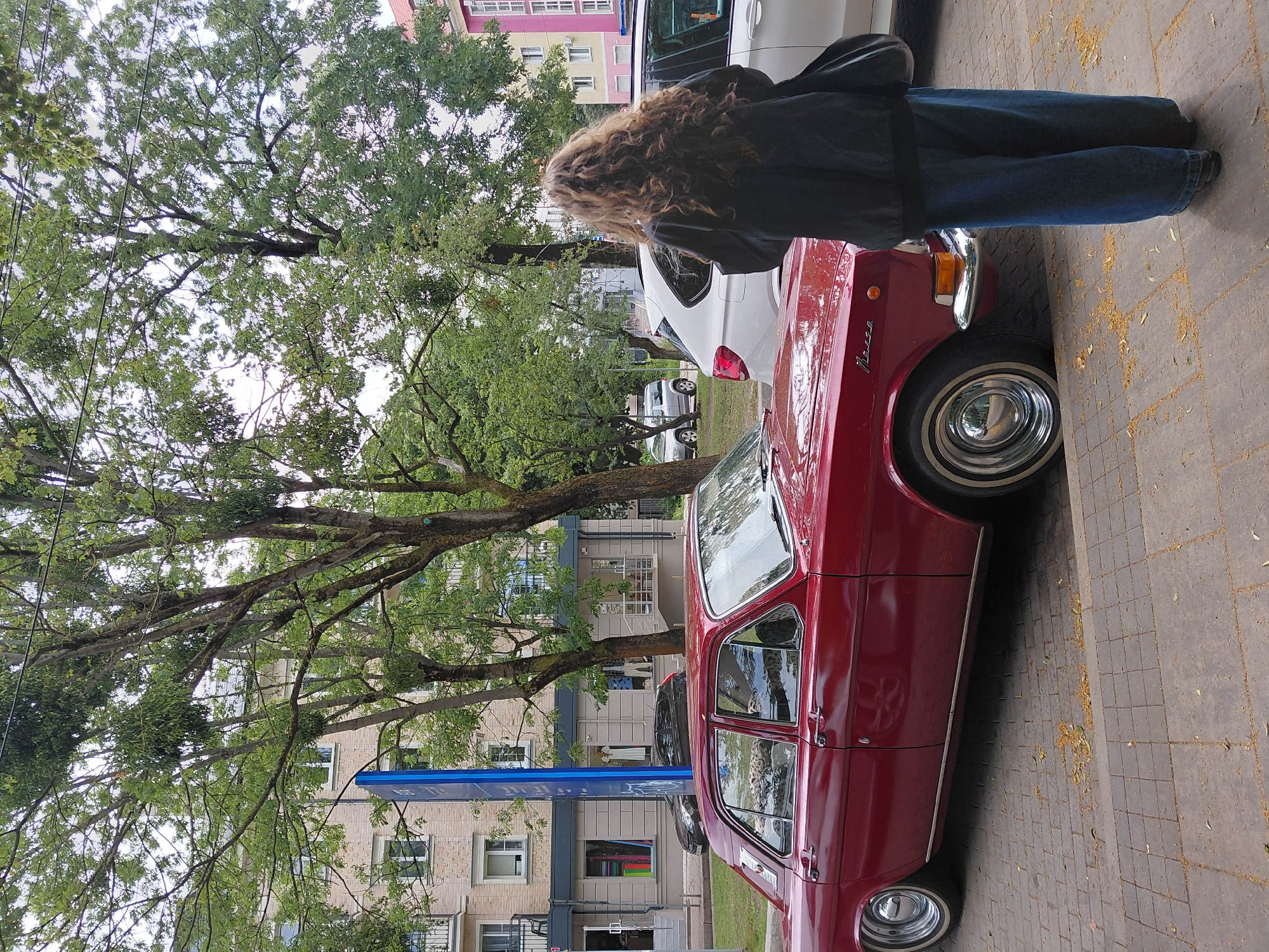 A person with long, curly brown hair, wearing a dark jacket and loose blue jeans, stands on a cobblestone sidewalk, facing away from the camera. The person is looking at a shiny, red, four-door sedan parked at the curb. The car appears to be a vintage Soviet-era model. Behind the car, there is a row of modern apartment buildings and shops with trees lining the street. The sky is overcast. The image appears to have been taken on a street in Kaliningrad, Russia.