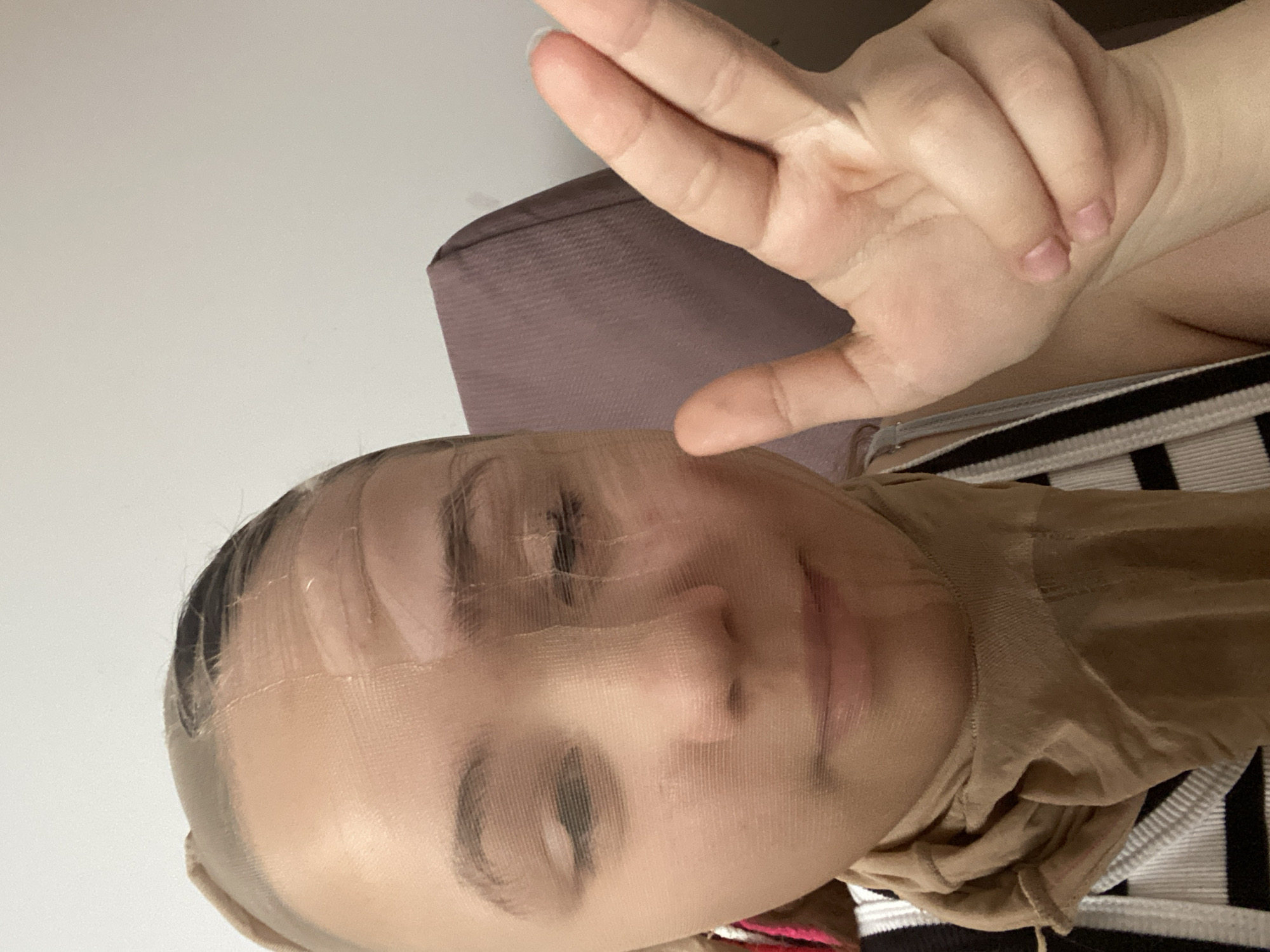 The image shows a person wearing a stocking over their head and face. The person is making a peace sign with their right hand. The background includes a white wall and a tan cushion. The person is wearing a striped shirt. The location is Unknown, Unknown.