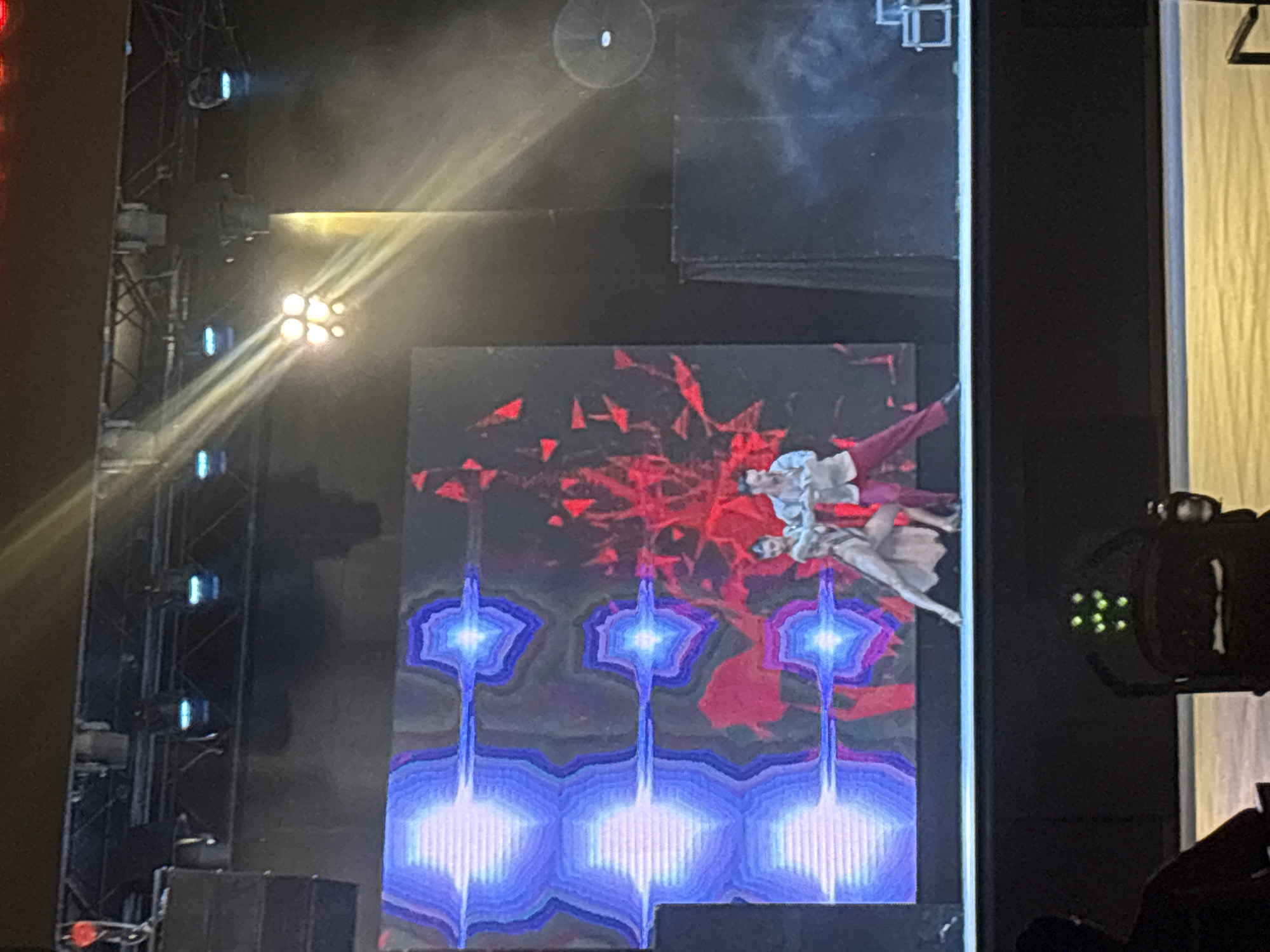 Two dancers, a male and a female, are performing on a stage in front of a large screen displaying abstract, colorful graphics. The male dancer is on the right, wearing a white shirt and red pants, and is positioned behind the female dancer. The female dancer is on the left, wearing a light-colored, flowing dress. The stage is lit by bright spotlights. In the foreground, a person is seated in a stroller or chair, facing away from the stage. The environment appears to be an indoor venue, possibly a theater or auditorium. The city and specific location are unknown.
