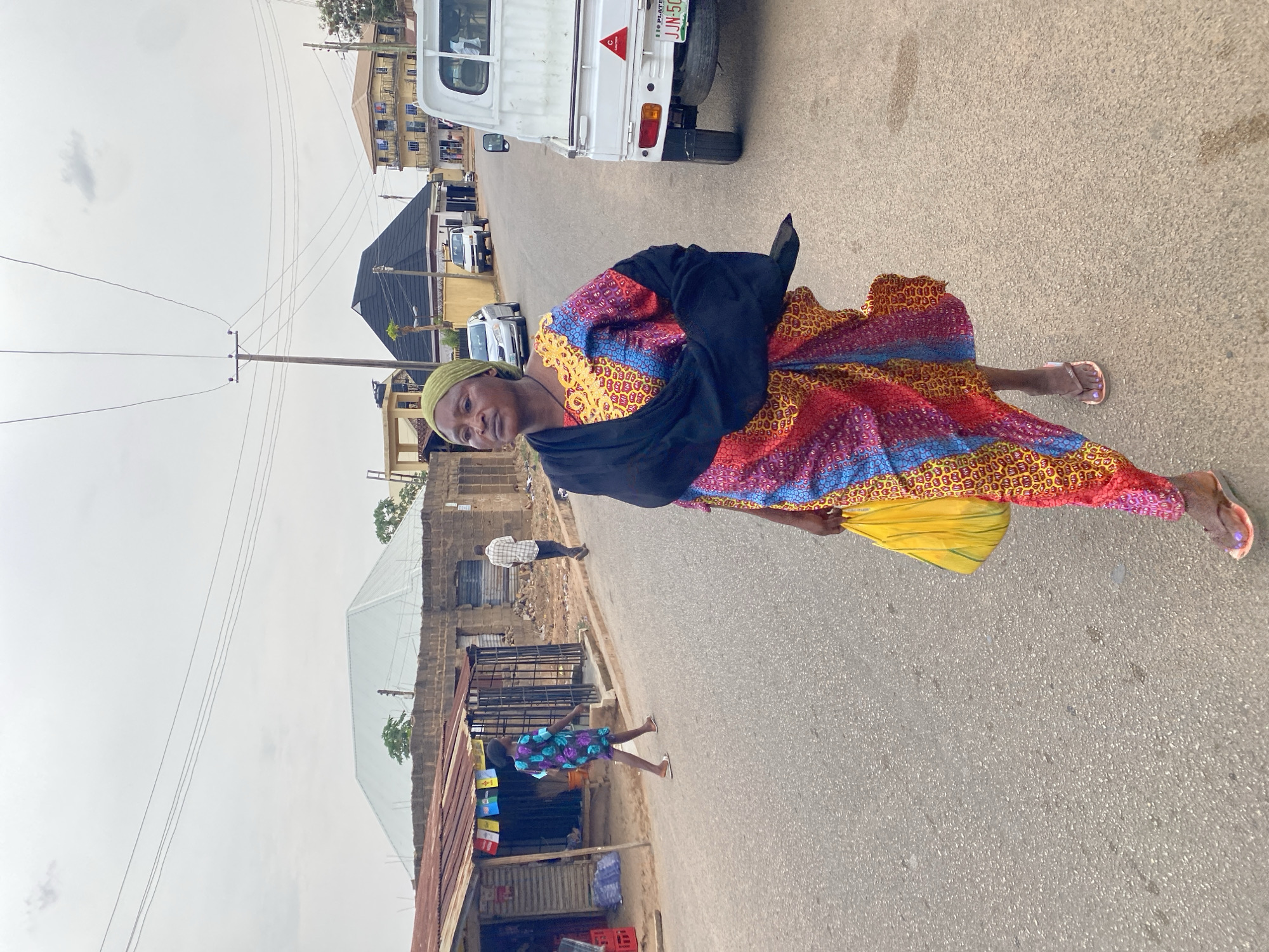 The image shows a woman standing on a paved road in Jos, Nigeria. She is wearing a colorful, patterned dress with a black shawl and a green headwrap. She holds a yellow bag. A white truck is parked on the road behind her. Buildings and utility poles line the street. In the distance, other vehicles are visible. A corrugated metal structure is present on the left side of the road. Another person is walking behind a child.