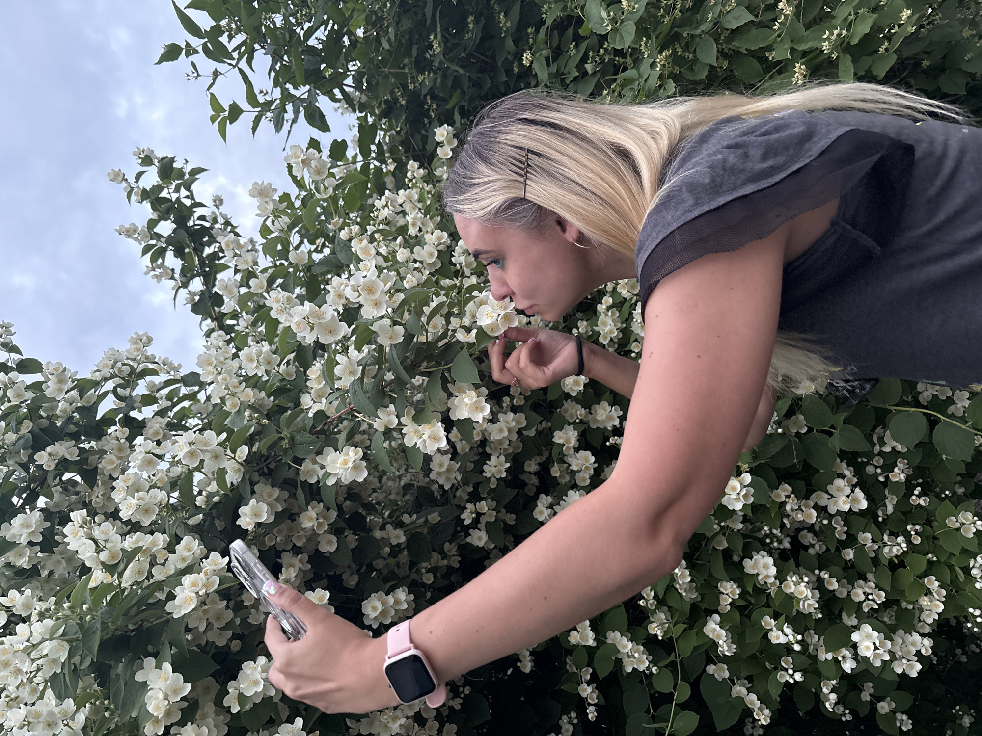 A young person with blonde hair and streaks of grey is standing in front of a bush with white flowers, taking a selfie with their phone. They are wearing a pink smartwatch on their left wrist. The background is a dense bush with small white flowers and green leaves. The sky above is cloudy. The image appears to be taken outdoors, possibly in a park or garden, given the presence of lush vegetation. The specific location in Chaikovsky, Russia, cannot be determined from the visual information alone.