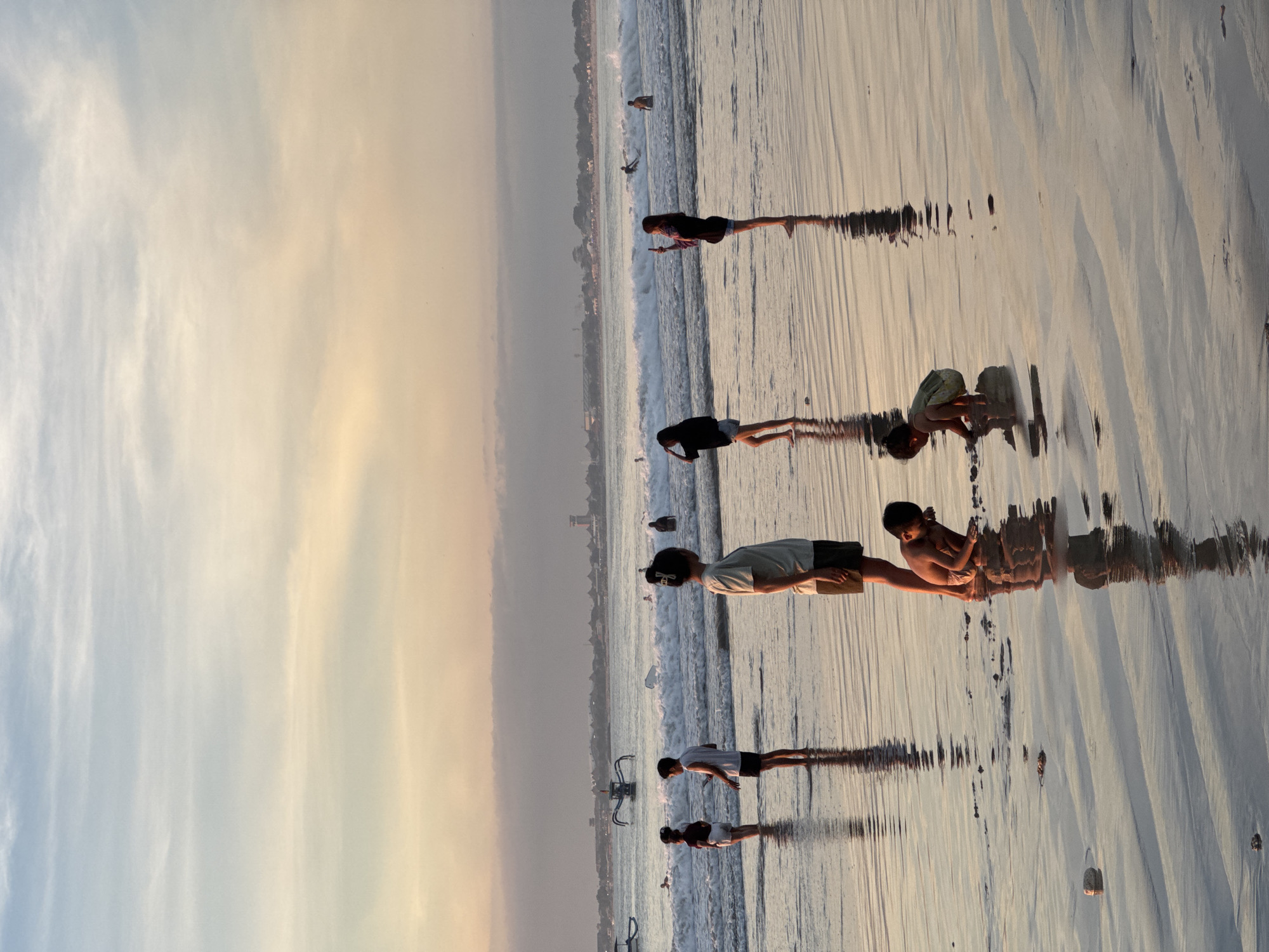 The image shows a scene on a beach in Kuta Selatan, Indonesia. The sky is filled with wispy clouds and a gradient of color. The beach stretches into the distance, with several people visible along the shoreline. Some people are standing near the water's edge, while others appear to be playing in the sand. Small waves are breaking along the shore, creating a pattern of ripples on the wet sand. There are boats and other objects scattered along the beach. The overall scene suggests a tranquil and serene atmosphere.