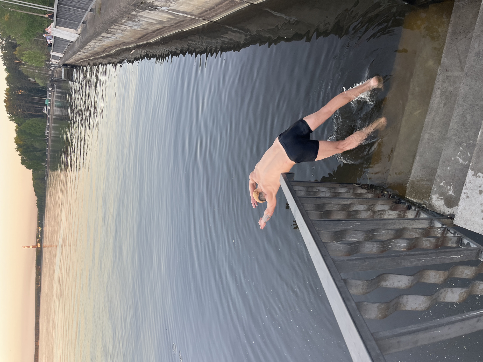A man is diving into water from a structure near a water body. The man is wearing dark-colored swim trunks and his body is angled, with arms outstretched toward the water and legs slightly raised. There are splashes of water visible around his legs, indicating recent entry or close proximity to the water's surface. The water has a slight ripple effect on its surface. To the right, there are concrete stairs or steps leading down to the water's edge. There's a metal railing or structure to the right of the frame.
In the background, the opposite shoreline shows a wooded area and a glimpse of structures that may be part of a city. The sky is pale and suggests it could be dusk or dawn. Based on the description, this could be taken in the city of Chaikovsky, Russia, considering it is a Russian city with a body of water nearby.