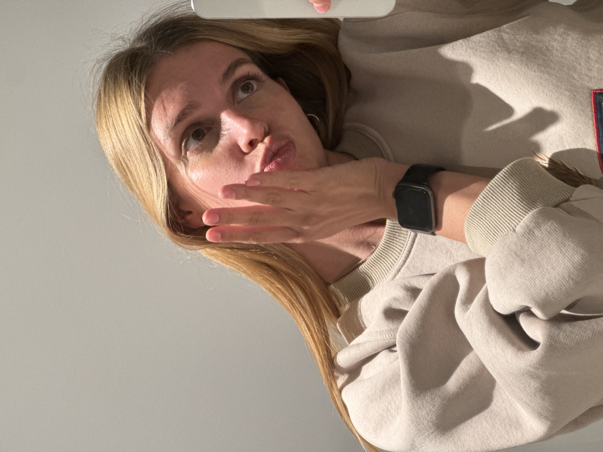 A woman with long blonde hair, wearing a beige sweatshirt and a black smartwatch, holds her hand up to her face with pursed lips, as if blowing a kiss. The photograph appears to be a selfie taken indoors. The background is a plain, light-colored wall. The exact location within Tbilisi, Georgia is indeterminable from the image.