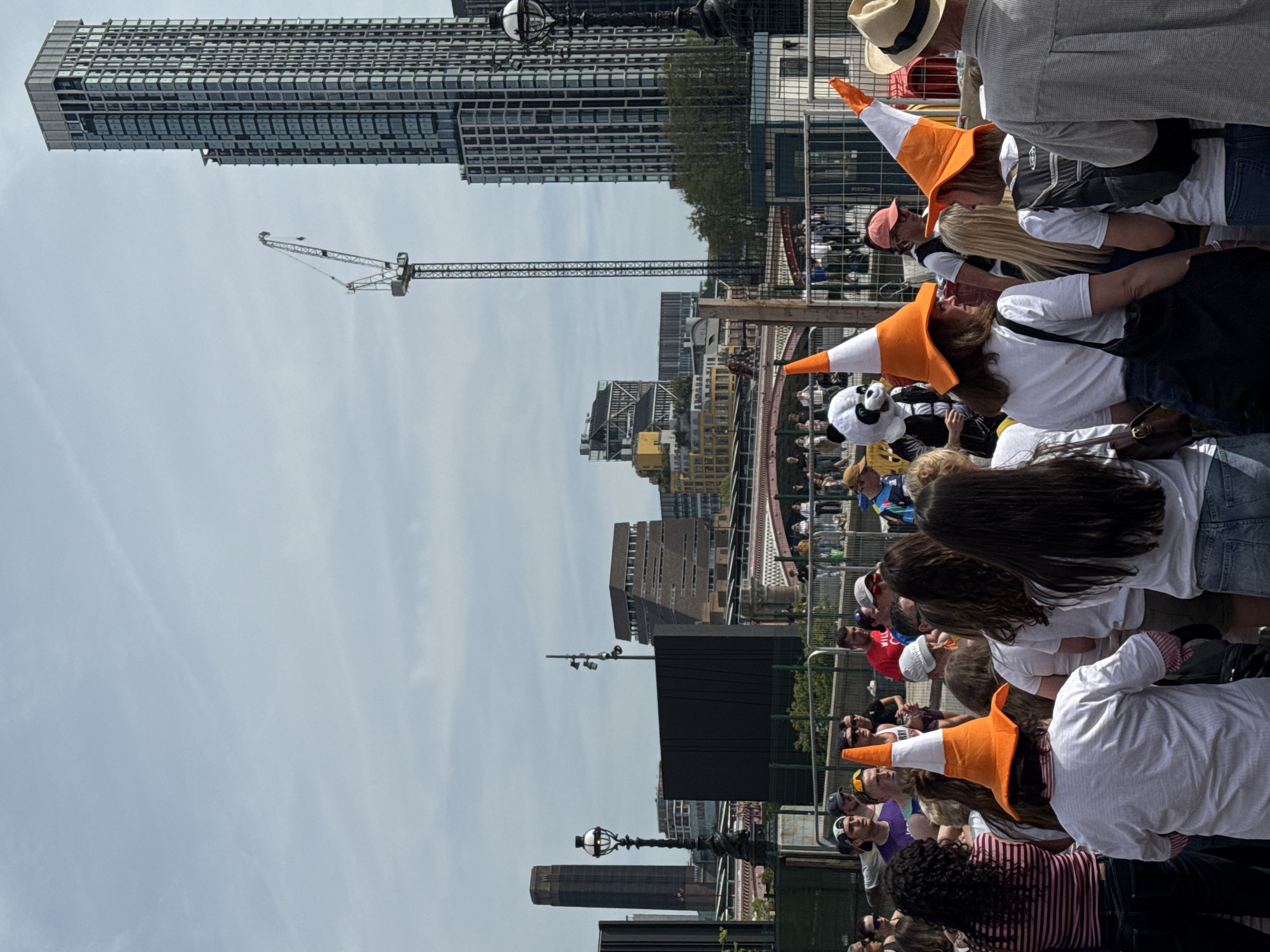 The image shows a crowd of people in London, United Kingdom. Many of the individuals are wearing orange and white traffic cone hats. The crowd is gathered near a barrier, and in the background, there are several buildings, including a tall skyscraper and the Tate Modern. A construction crane is also visible in the distance. The day appears bright with a cloudy sky.