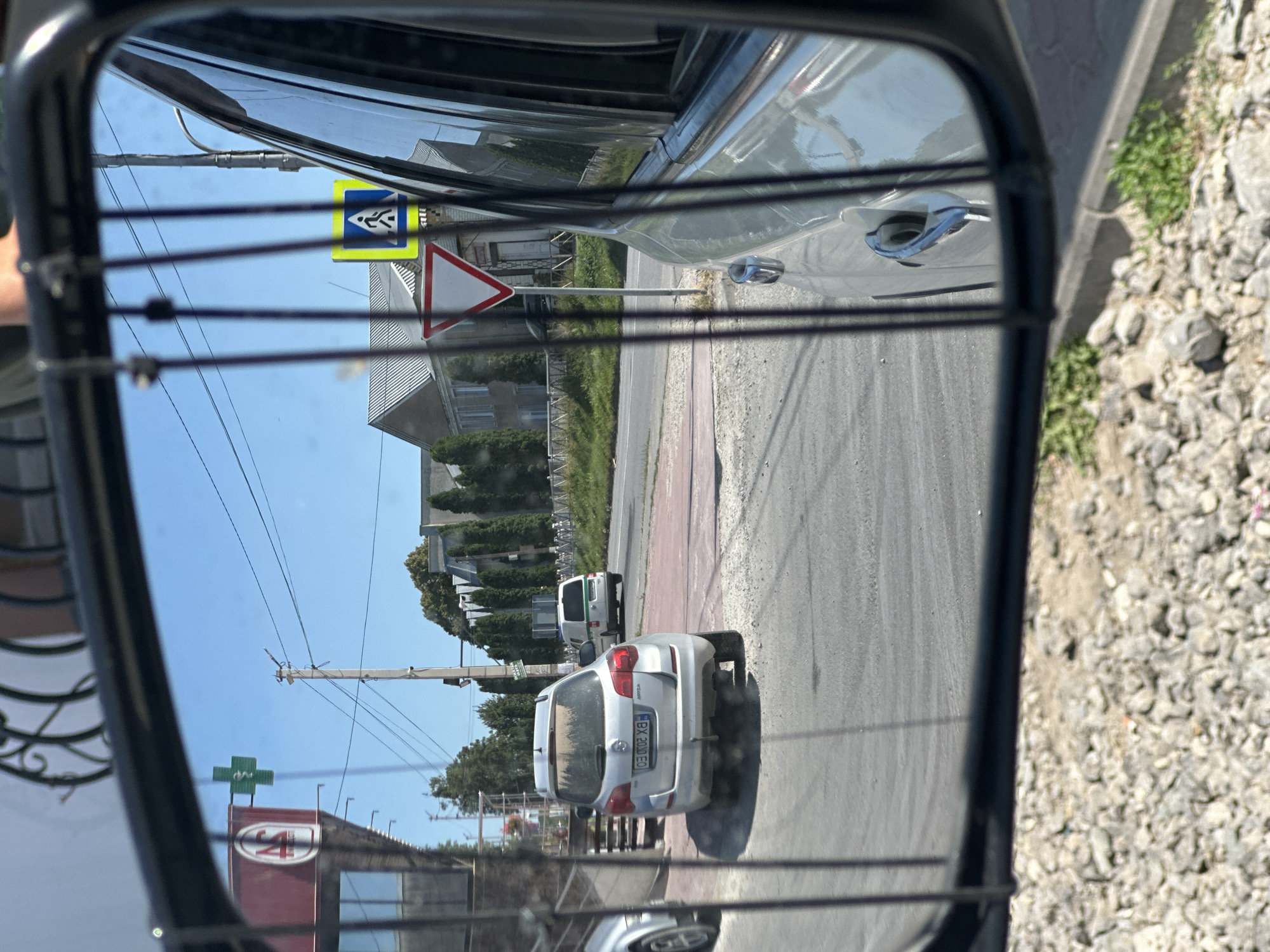 The image is a reflection in a car's side-view mirror, showing a street scene in Shatava, Ukraine. In the foreground of the reflection, a silver SUV is parked. Beyond the SUV, a road is visible with a pedestrian crossing sign and a give way sign. A white van is driving away on the road. Buildings and trees are visible in the background. A green cross sign on a red building is visible on the left side of the reflection. The sky is clear and blue. The reflection is partially obscured by vertical black bars, which appear to be part of the mirror's housing.