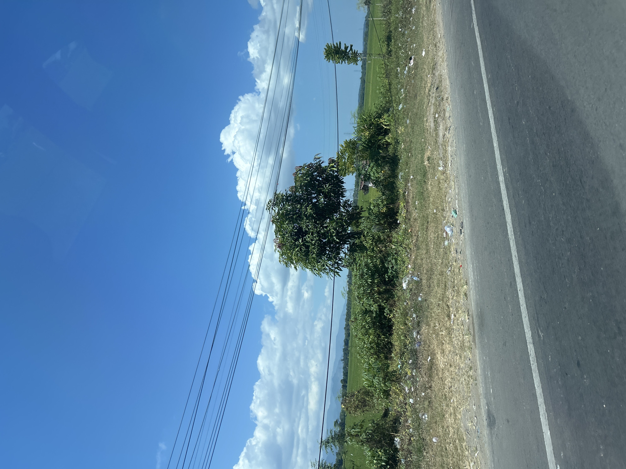 The image captures a roadside scene in Kalabbirang, Indonesia, under a clear blue sky dotted with fluffy white clouds. A paved road with a white dividing line runs through the foreground, leading the eye towards a patch of dry, grassy land cluttered with white and translucent plastic debris. Beyond this, a vibrant green rice paddy stretches out, bordered by lush vegetation and scattered trees. In the mid-ground, a medium-sized tree with dark green foliage stands prominently. In the distance, a range of mountains forms a hazy blue backdrop against the horizon. Several electrical wires are strung across the top of the frame, supported by unseen poles. The overall impression is of a rural landscape, likely a roadside view from a moving vehicle, with evidence of human presence in the form of the road, wires, and discarded litter.
