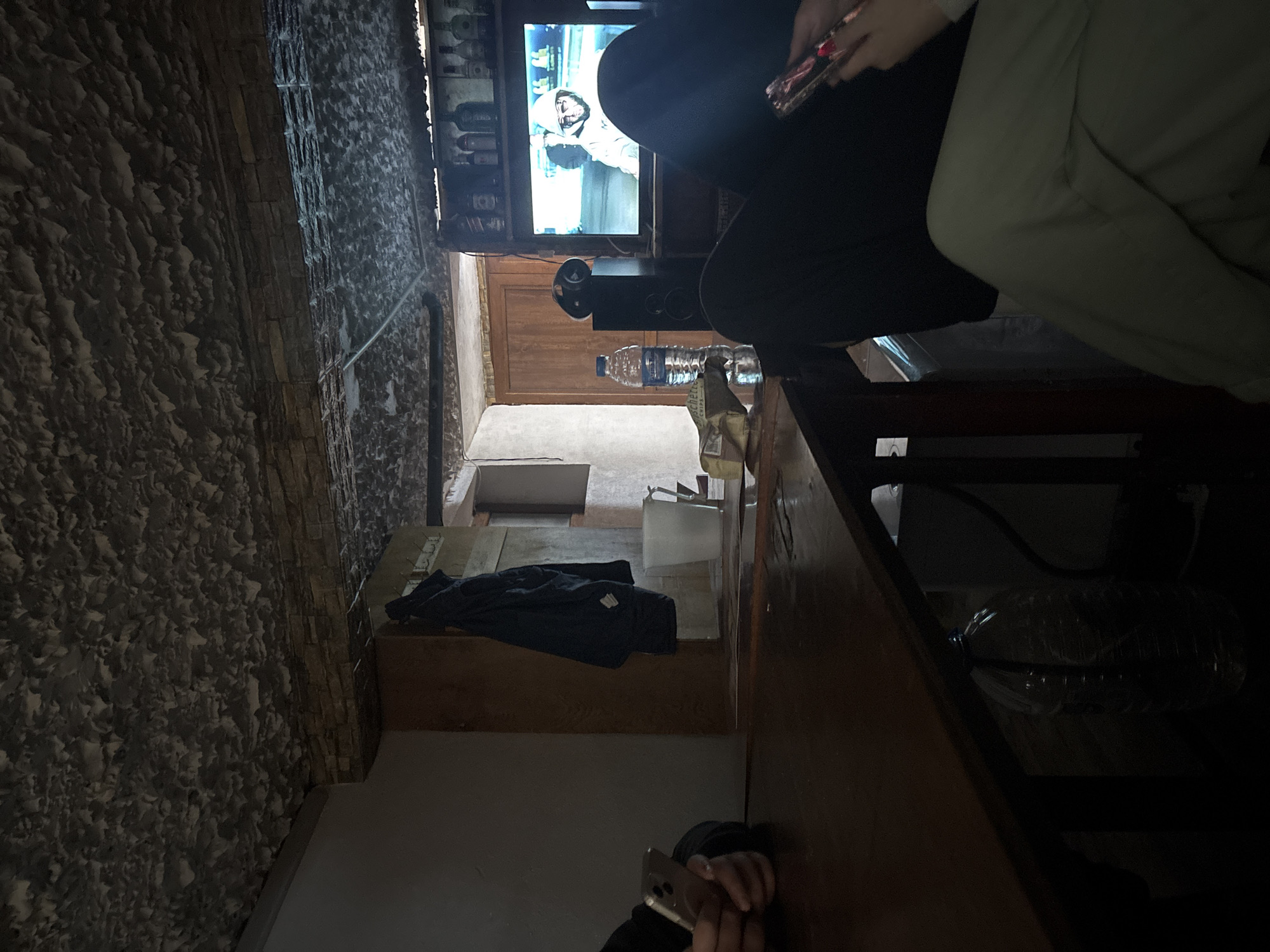 The image shows an indoor scene in Lehchevo, Bulgaria. The camera is positioned at an oblique angle. A person is sitting in the foreground, holding a phone in their right hand. Another person is sitting closer to the television, holding a phone. A television shows a scene with a person wearing a white garment. A bottle of water sits on the table. A blue jacket lies on a shelf. The walls are textured, and there appears to be a doorway in the background.