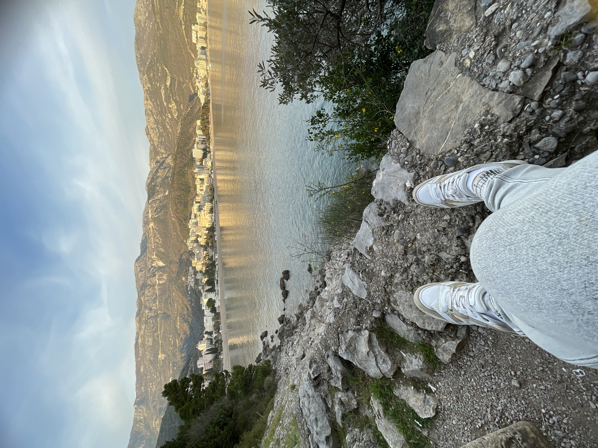 A first-person perspective photograph depicts the lower body of an individual seated on a rocky, uneven incline within Boreti, Montenegro. The person's legs are extended forward, clad in light grey denim jeans and white, grey, and gold athletic sneakers. The immediate foreground consists of a coarse gravel path and large, irregular grey rocks, interspersed with sparse green vegetation, including small bushes, some bearing yellow flowers, along the edge of the slope.
The view extends over a calm body of water, likely a bay, which reflects the sky and distant structures. Across this water, a densely developed coastal city, consistent with areas of the Budva Riviera, is visible. This city features numerous multi-story, predominantly light-colored buildings lining a sandy beach. Behind the urban sprawl, rugged mountains with visible winding roads rise steeply, forming the distant backdrop. The sky above is clear blue with wispy white clouds. The scene is illuminated by natural, warm-toned light, suggesting either early morning or late afternoon.