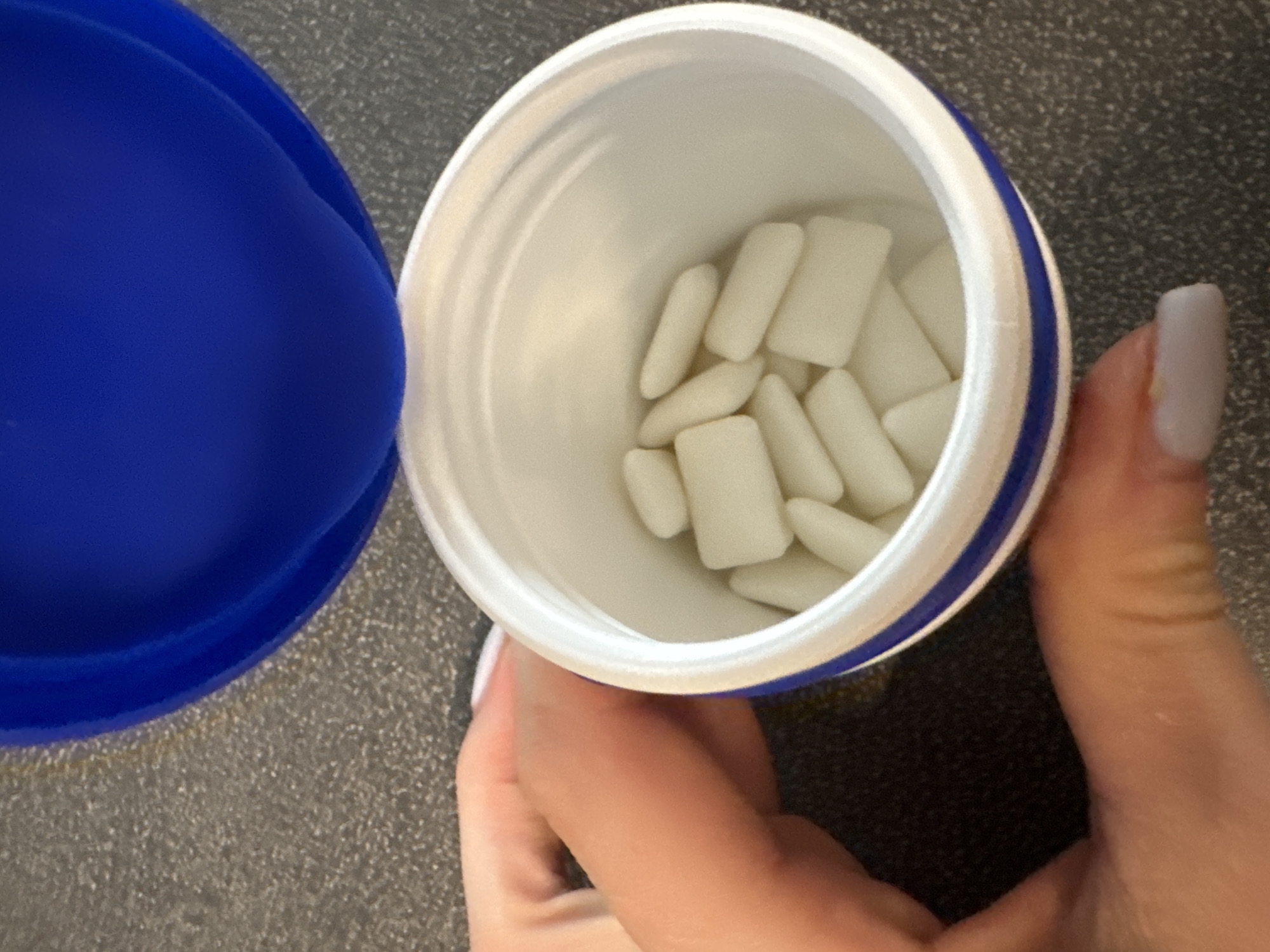 The image shows an open container of white, rectangular-shaped chewing gum pieces. The container is white with a blue band around the rim and is partially filled with the gum. To the left of the container is a blue lid, also open. A person's hand, with a light purple nail polish on the visible fingernail, is holding the container from the right. The background is a dark, speckled surface, possibly a countertop. The lighting suggests it is daytime, and the image appears to be taken indoors. No text is visible in the image. The scene is mundane, focusing on the chewing gum container.
