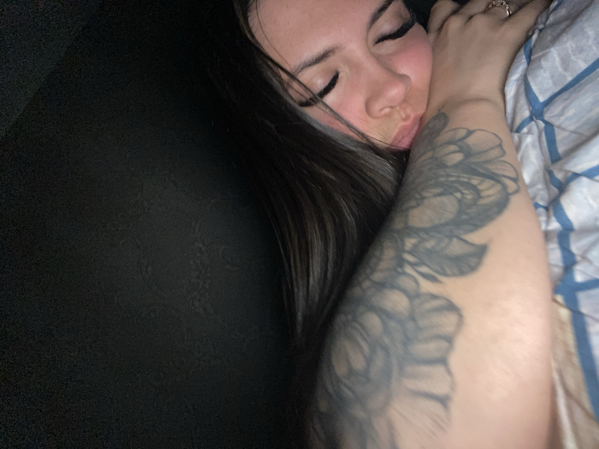 An individual, likely female, is depicted sleeping in an indoor environment in the city of Chaikovsky, Russia. The person's face is partially obscured as their head rests on their left arm, with eyes closed and dark hair fanned out. Eyeliner is visible along the upper eyelids. A prominent tattoo featuring dark, wing-like or feather-like patterns covers a significant portion of the left arm. A ring with multiple bands is present on the ring finger of the visible right hand. The individual is lying on a bed covered with a white comforter or sheet patterned with blue and grey squares. The background above the person is uniformly dark.