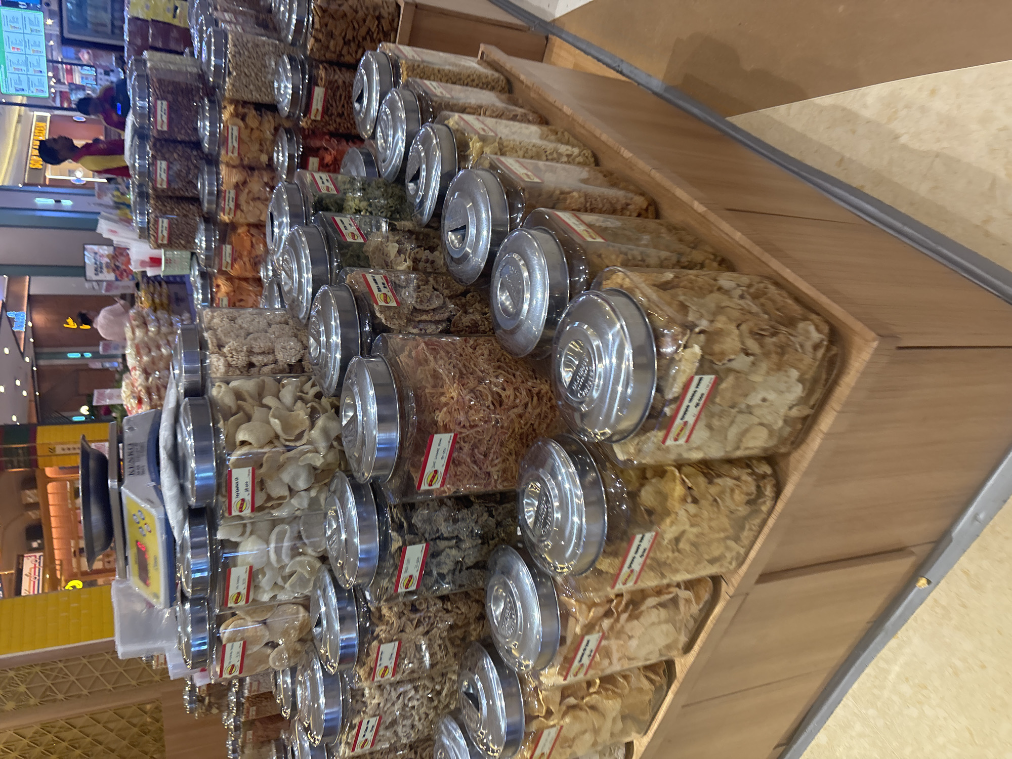 This is a medium shot of a snack stall in a shopping mall in North Jakarta, Indonesia. The stall features rows of large glass jars filled with various snacks, including chips, nuts, and dried seafood. The jars are topped with silver lids and have small red and yellow labels. In the background, shoppers can be seen browsing at other stalls. A digital scale sits on a shelf behind some of the jars, suggesting that the snacks are sold by weight. The flooring is a mix of beige tiles and wood.
