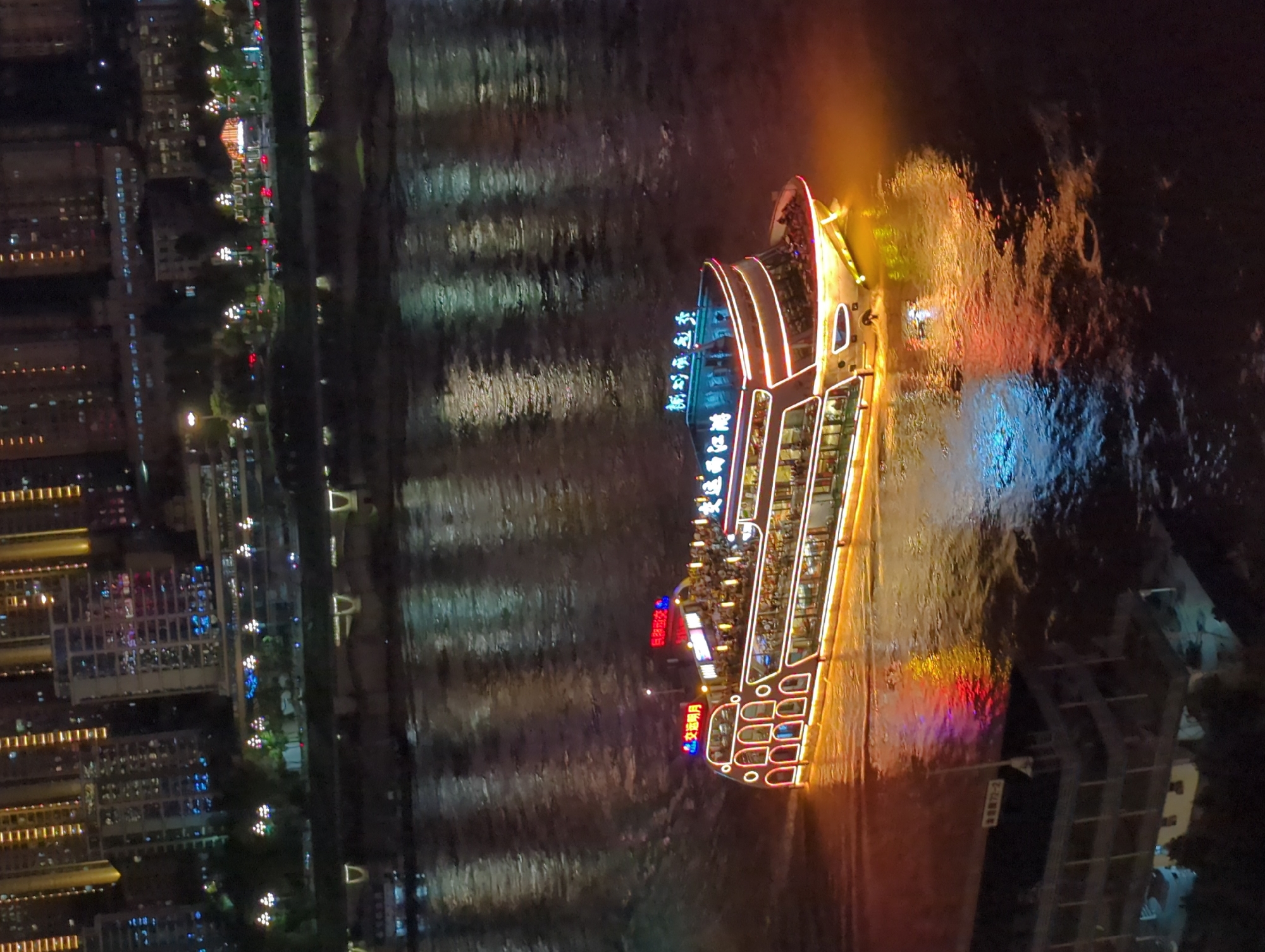 The image shows a brightly lit, multi-decked passenger boat navigating a dark body of water at night. The boat is adorned with numerous lights, outlining its structure and reflecting on the water's surface, creating a vibrant spectacle of color. The water itself is dark, with shimmering reflections of the city lights and the boat's illumination.
In the background, a city skyline rises, characterized by tall buildings with rows of illuminated windows. The buildings are a mixture of modern architecture, with some featuring vertical lighting accents. The city lights are varied, including white, yellow, blue, and red hues, suggesting a bustling urban environment. The lights of the boat are primarily white and red, with some yellow and blue accents, and they cast a warm glow on the water directly around it.
The boat appears to be crowded with people on its upper decks, visible as silhouettes against the bright lights. The boat's hull is white, with multiple rows of windows. The name of the boat is visible in illuminated Chinese characters on the top deck. The overall scene suggests a nighttime cruise or ferry operation on a river or bay in a major Chinese city, likely Chongqing's Yuzhong District, given the presence of illuminated buildings and waterways typical of the area. The water appears to be slightly choppy, with ripples and waves visible.