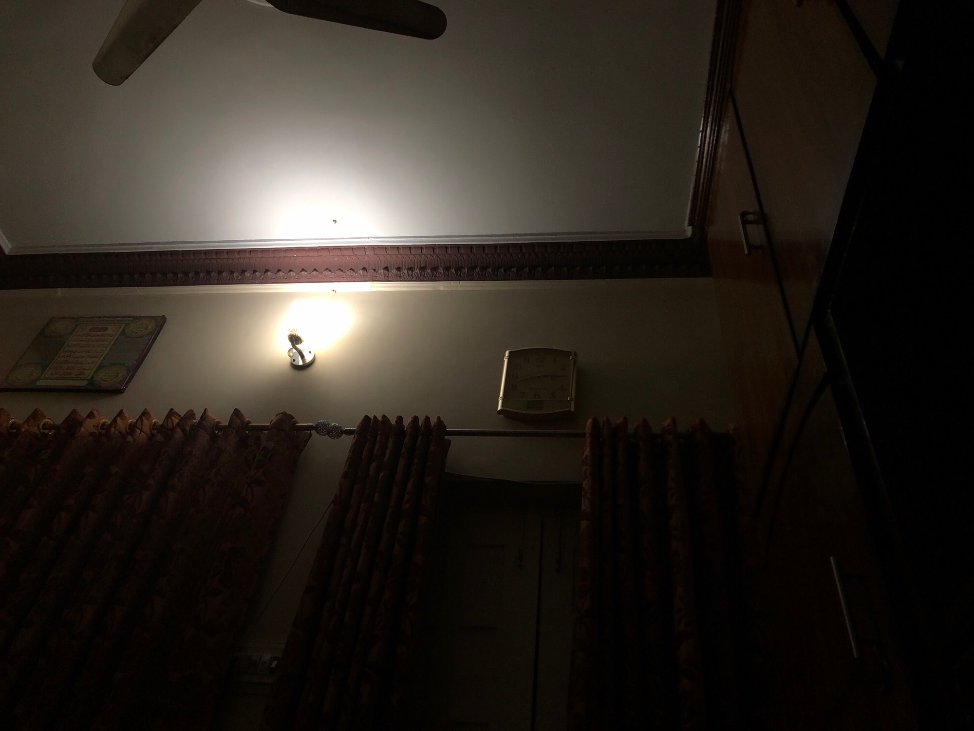The image shows a dimly lit room, likely in Larkana, Pakistan, with a ceiling fan visible at the top. On the wall, a light fixture illuminates the space, casting a glow on a decorative border and a framed artwork. To the right, a clock is mounted on the wall above a set of heavy, patterned curtains. Further to the right, a tall, dark cabinet or wardrobe dominates the scene, its surface reflecting some light. The overall impression is of a residential interior, possibly captured at night or with low lighting.