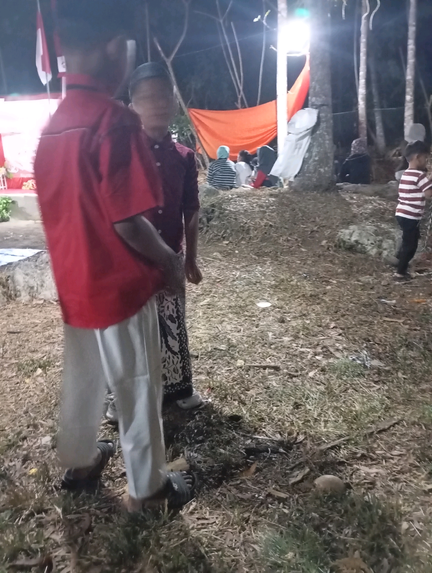 The image depicts an outdoor scene at night, likely in Ketepung, Indonesia, with a gathering of people. The foreground features two young males. The individual on the left wears a red long-sleeved shirt and light-colored pants, with dark sandals. The person to their right wears a darker, possibly maroon, long-sleeved shirt and a patterned sarong, also with dark sandals. In the background, other individuals are visible, some seated and some standing. There is an orange tarp and a white tarp draped over trees and a structure, illuminated by bright lights. The ground is covered with dry leaves and grass. The scene suggests a community event or informal gathering taking place outdoors after dark.