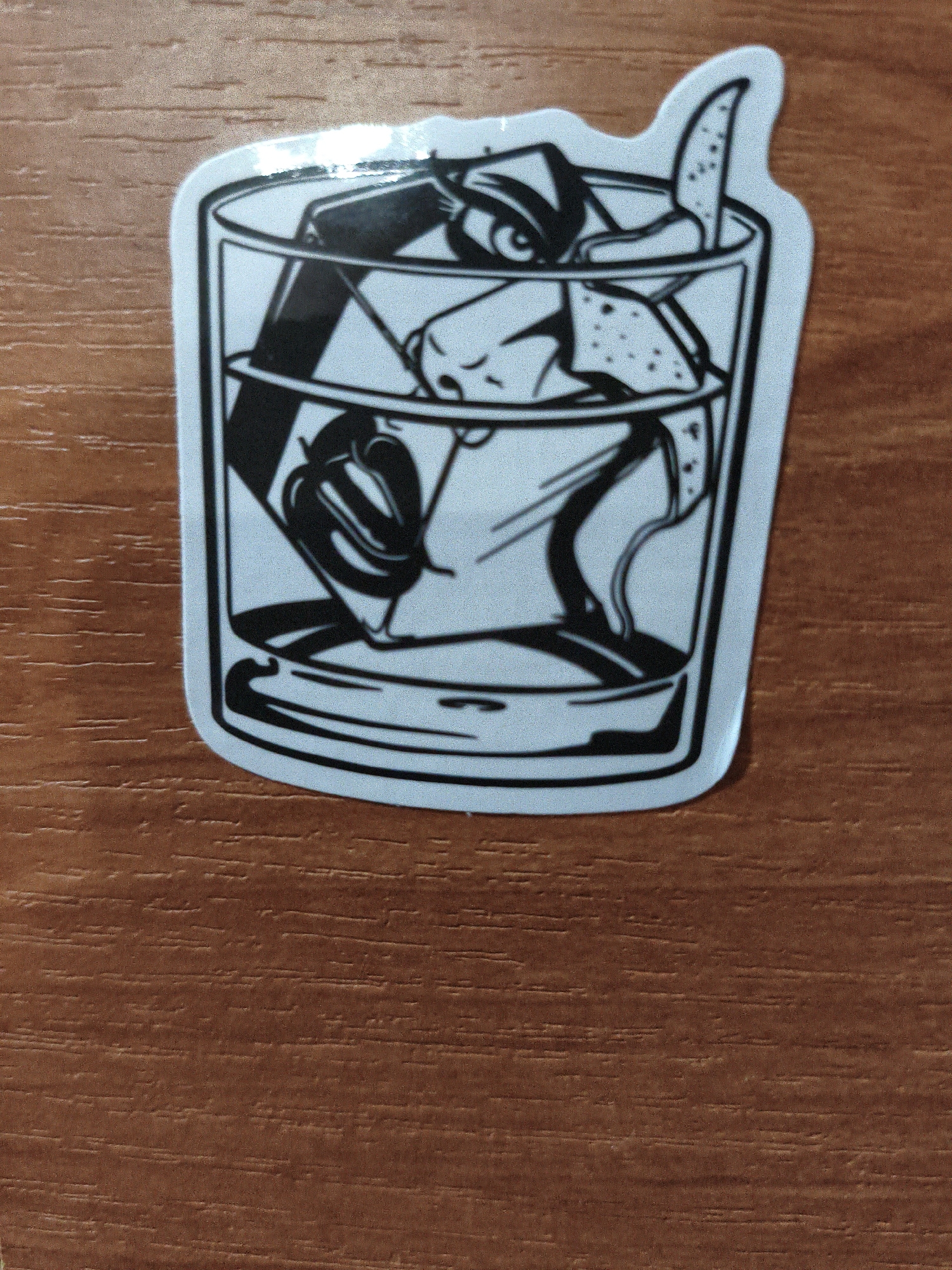 A black and white sticker depicting a close-up of a glass filled with ice and a drink is shown on a wooden surface. The sticker has a white border and a slightly irregular, cut-out shape. The drawing within the sticker is stylized, featuring a face partially submerged in the drink, with eyes visible above the liquid. The background of the image is a brown wooden surface with a visible wood grain pattern. This image does not provide any information related to the city of Lyubertsy, Russia, or any specific location.