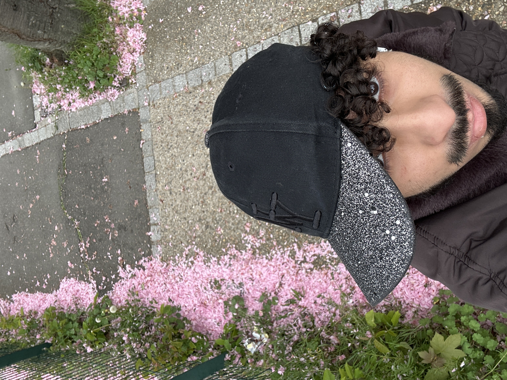 A person is oriented in an upright position, wearing a black baseball cap with a glittery bill and a dark jacket. The person has curly hair, a beard, and visible skin. The person is on a paved surface with pink flower petals scattered around. The petals appear to be concentrated at the base of some vegetation, which includes green grass. The image appears to be taken from a high angle. The location is possibly in Orsay, France, based on the type of paving stones and flowers commonly found in that region.