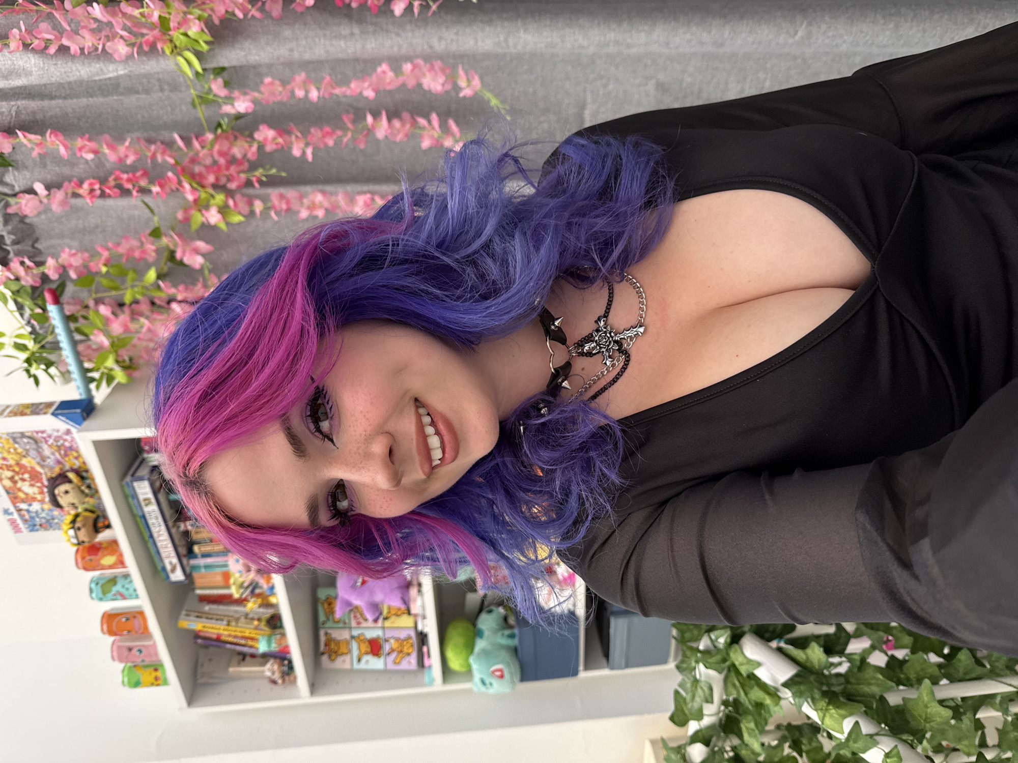 A young woman with vibrant, long pink and purple hair is featured in this portrait, smiling directly at the camera. She is wearing a black, long-sleeved top with a low neckline, and a distinctive black choker necklace with spikes and a silver rose or cross pendant. Her makeup includes winged eyeliner and freckles across her nose.
The image is taken indoors and is oriented 90 degrees counter-clockwise. The background reveals a white shelving unit stocked with various items, including books, small figurines resembling characters like Naruto, Pikachu, and Winnie the Pooh, and what appears to be a puzzle box. Decorative elements include pink floral garlands draped against a grey backdrop and green ivy garlands visible in the lower right portion of the frame. The woman is actively posing for the photograph, displaying a friendly and happy expression. The indoor lighting suggests the photo was taken during the day. No legible text is visible within the image. This scene is situated in the City of Zagreb, Croatia.