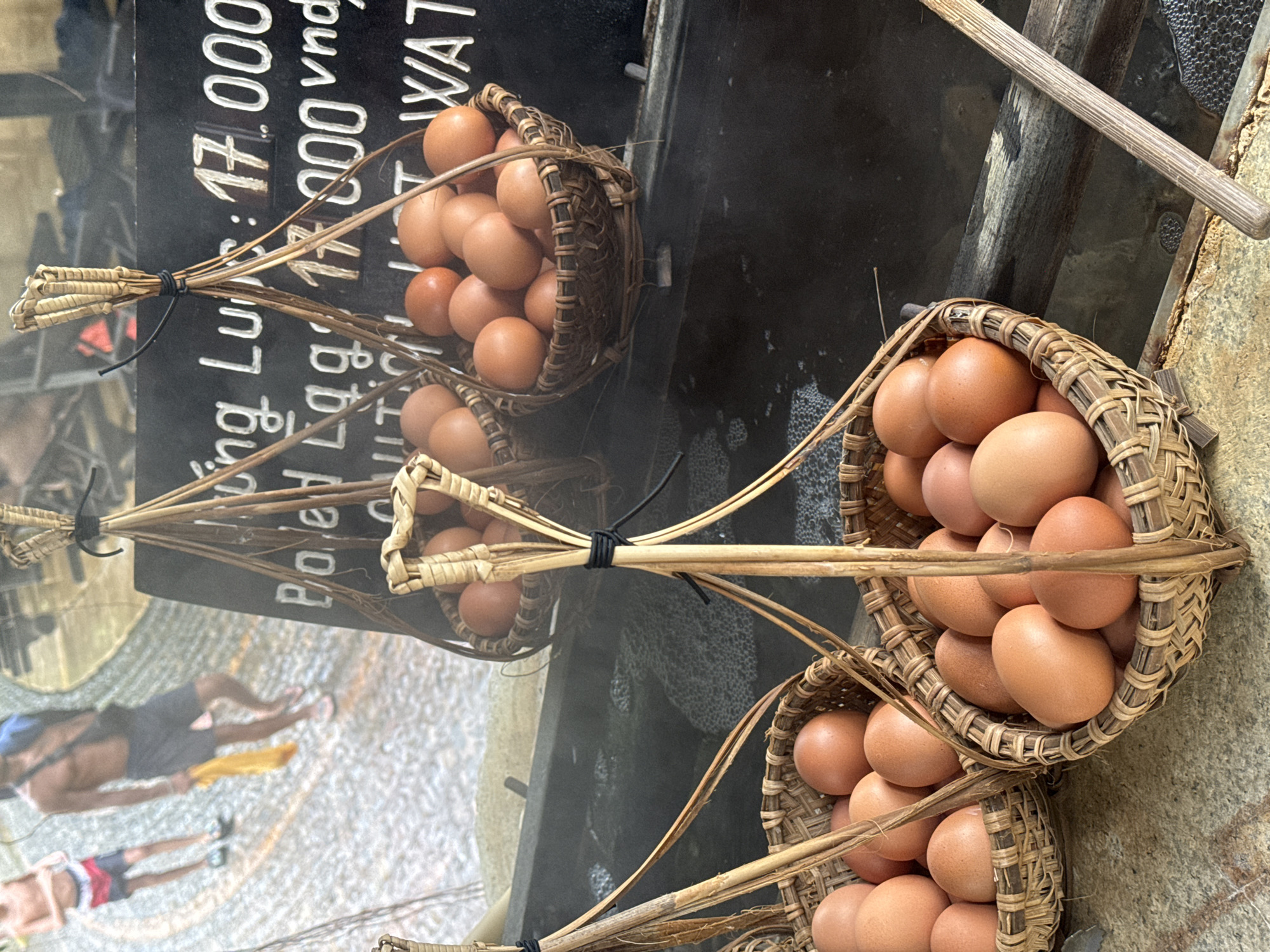 In Nha Trang, Vietnam, an outdoor scene showcases baskets filled with brown eggs. In the foreground, two woven baskets brim with eggs, with one basket featuring a prominent, large egg in the center. A wooden stick, likely used for handling, lies horizontally in the lower right corner. In the background, two men, one wearing red and blue swim trunks and the other dark shorts, walk on a textured surface. A black sign with white lettering, partially obscured by hanging baskets and eggs, displays text that appears to include "Boiled Eggs" and currency symbols. The overall setting suggests a market or food stall where boiled eggs are being sold.