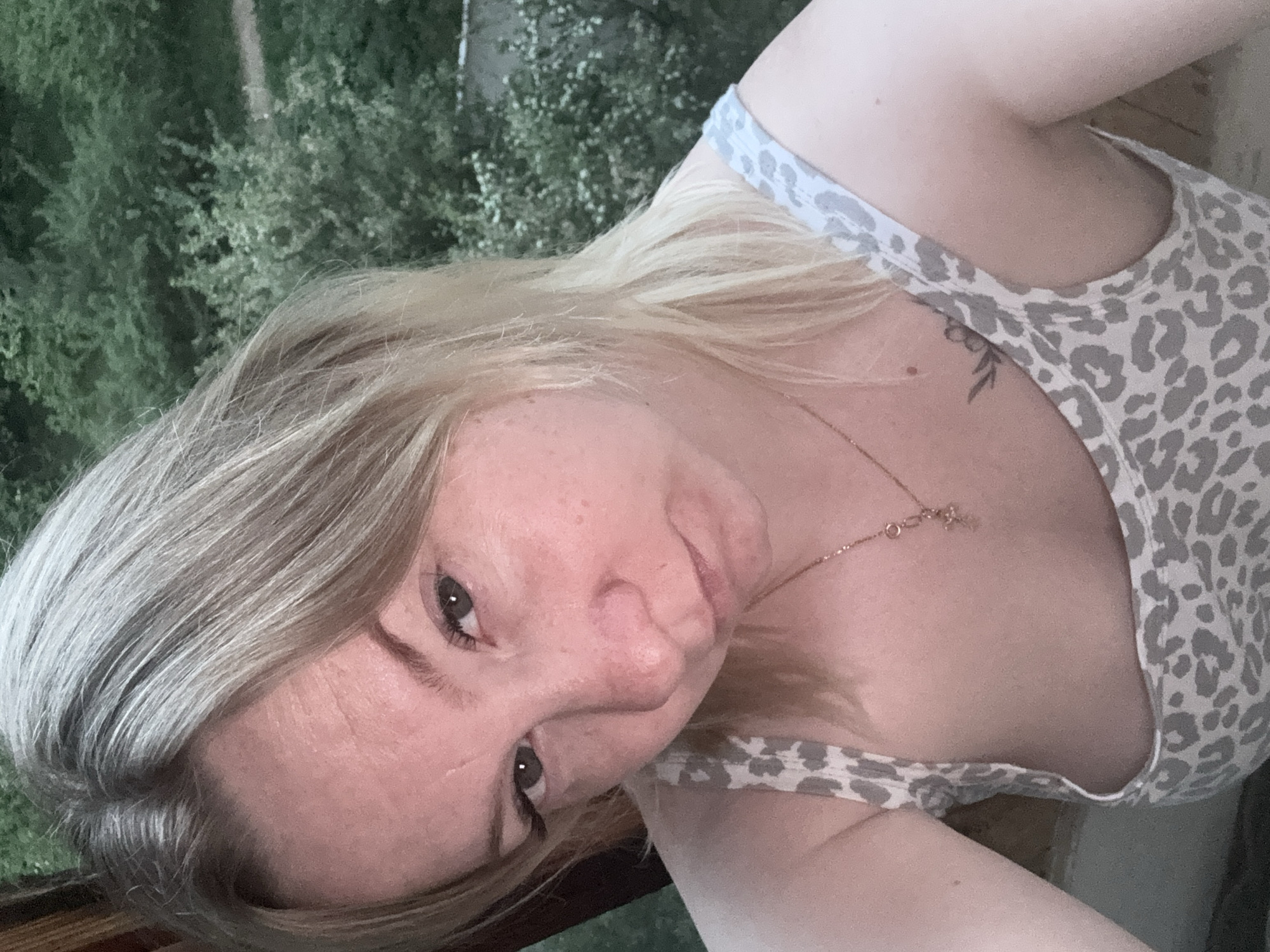 A woman with blonde hair and fair skin is photographed in an indoor setting against a background of greenery, possibly foliage viewed through a window. She is wearing a light-colored animal print tank top and a delicate gold necklace. A small tattoo is visible on her upper chest. The photograph is slightly tilted. No specific details in the image indicate that it was taken in Chaikovsky, Russia.