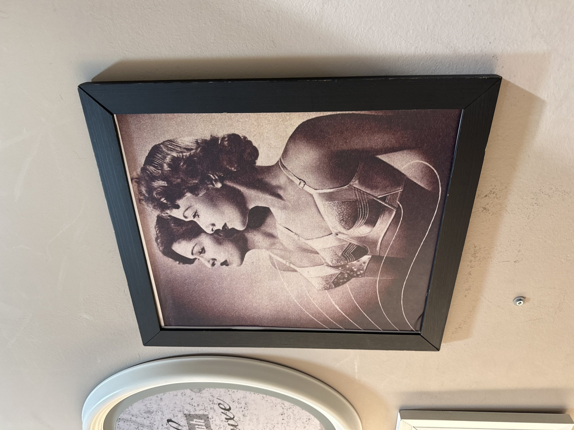 The image shows a framed picture hanging on a wall. The picture is a sepia-toned image of two women in lingerie. The frame is black and appears to be made of wood. Below the framed picture, a portion of another white framed object is visible. The wall is light beige. A screw is seen slightly to the right of the upper right corner of the frame. It is not possible to identify a location in Sofia, Bulgaria, from this image.