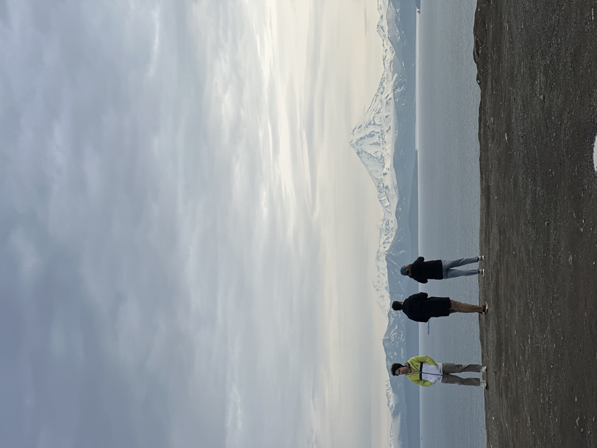 Three individuals stand on a dark, rocky cliff overlooking a vast body of water. The person on the left, facing left, wears a light green and white jacket and tan pants. The person in the middle, facing away from the camera, wears a black coat and brown pants. The person on the right, also facing away, wears a dark jacket and blue jeans, with a blue baseball cap. In the background, a majestic mountain range, capped with snow, stretches across the horizon. The water is calm and reflects the overcast, cloudy sky. The scene appears to be a viewpoint, possibly a scenic overlook, offering a panoramic view of the natural landscape. Based on the distinctive snow-capped volcanic mountains and the general geographic context, this location is likely the viewpoint overlooking Avacha Bay and the surrounding volcanoes near Petropavlovsk-Kamchatsky, Russia, such as the Koryaksky and Avachinsky volcanoes.