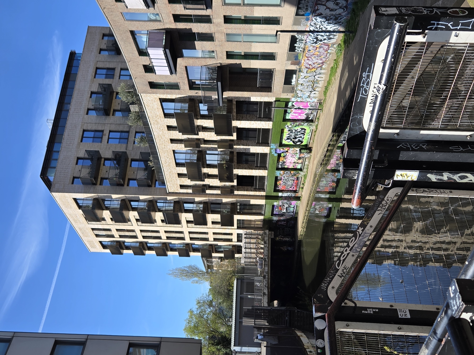 The photograph shows a canal in London, United Kingdom, with an apartment building rising on the left side of the frame. The canal water reflects the building and a graffiti-covered wall running alongside the water. A structure made of metal grates and tubes, possibly part of a boat or canal infrastructure, is prominently displayed in the foreground, with graffiti markings on its surface. The sky is a clear blue.