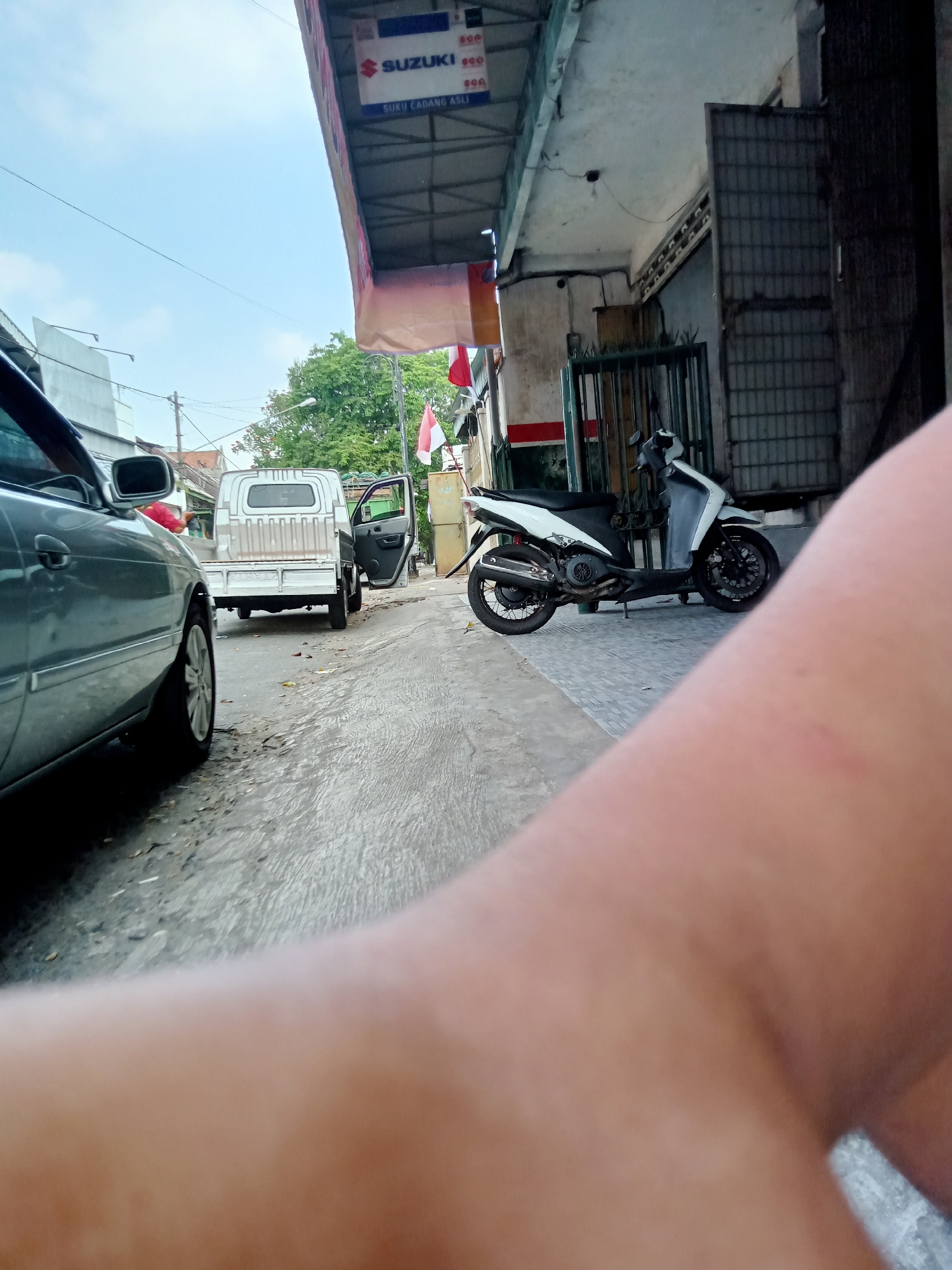 The image depicts a street scene in Tulungagung, Indonesia. In the foreground, a person's arm is partially visible, blurred by the camera's close proximity. To the left, a silver car is parked. Further down the street, a white pickup truck with its driver's side door open is stationary. On the right side of the street, a white scooter is parked. Above the scooter, a store canopy with "SUZUKI" branding and the text "SUKU CADANG ASLI" (Genuine Spare Parts) is visible. An Indonesian flag, red and white, is also displayed. The environment suggests an urban setting with buildings and overhead wires. The ground appears to be paved and somewhat wet, with scattered debris. The sky is overcast.