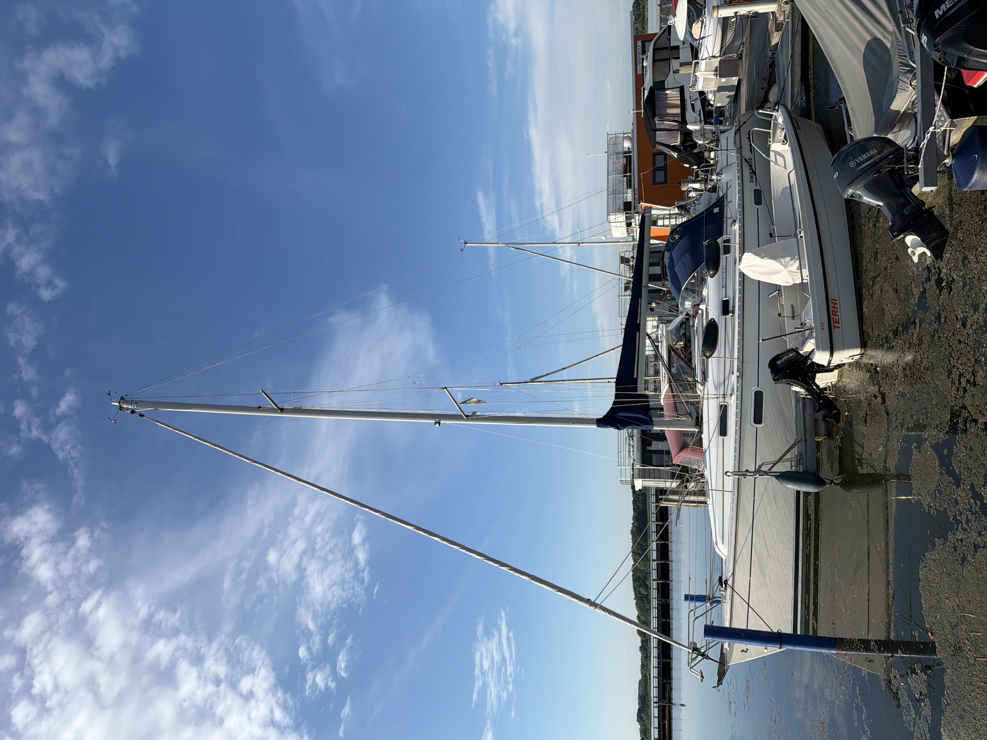The image shows several sailboats docked at a harbor. The boats are white with blue accents and have tall masts. Some of the boats have outboard motors. The water is calm and reflects the blue sky, which is dotted with white clouds. The sun appears to be shining, suggesting it is daytime. The scene is set in a harbor with a wooden dock and a shoreline with trees in the distance. The overall atmosphere is peaceful and serene.
Visible text on the boats includes "TERHI 450", "BENETEAU", "Diega and", and "YAMAHA". The location context is Ribnitz-Damgarten, Germany. There are no people visible in the image. The activity taking place is the docking of sailboats, suggesting a marina or harbor setting. The time of day is likely daytime, given the bright sunlight and blue sky. The weather appears to be fair.