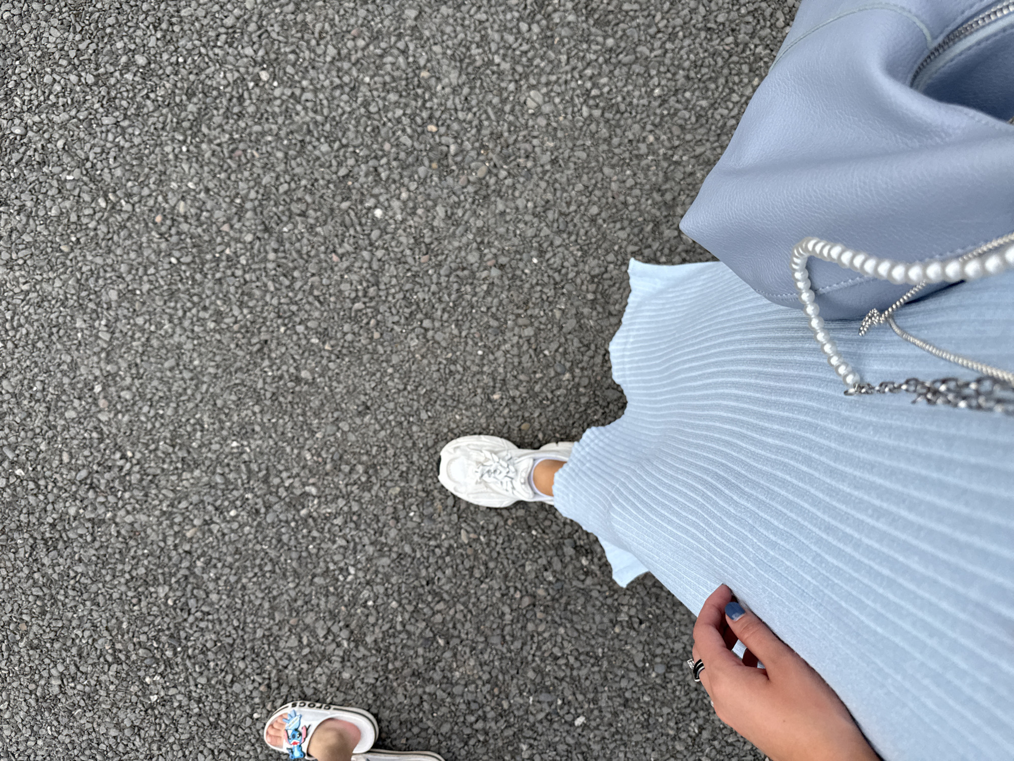 The image shows a low-angle perspective of a person walking on asphalt, with another person's footwear visible in the upper left corner. The primary subject is wearing a light blue, pleated dress and white sneakers. Their hand, adorned with rings and blue nail polish, rests on the dress. A light blue handbag with a pearl strap is also visible. The environment appears to be an outdoor urban setting, possibly a street or sidewalk in Bely Yar, Russia, based on the provided context. The asphalt surface suggests a developed area.