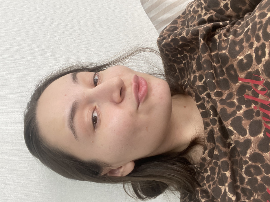 An indoor portrait depicts a single individual, a female with medium-brown hair, dark eyes, and a fair to medium skin tone exhibiting minor blemishes. She is looking directly at the camera with her lips pursed in a kissing or puckered expression, accompanied by a slight upward curve at the corners of her eyes. Her hair is styled down, partially covering her left ear and framing her face. She is wearing a dark brown and tan leopard print top, with partial red lettering visible on the chest that appears to spell "wild." The background consists of a plain, light-colored wall with a subtle vertical textured pattern. To the subject's right, a striped object, possibly a pillow or bedding, is partially visible. The exact location within the city of Соболиха, Russia, cannot be determined from the visual information provided.