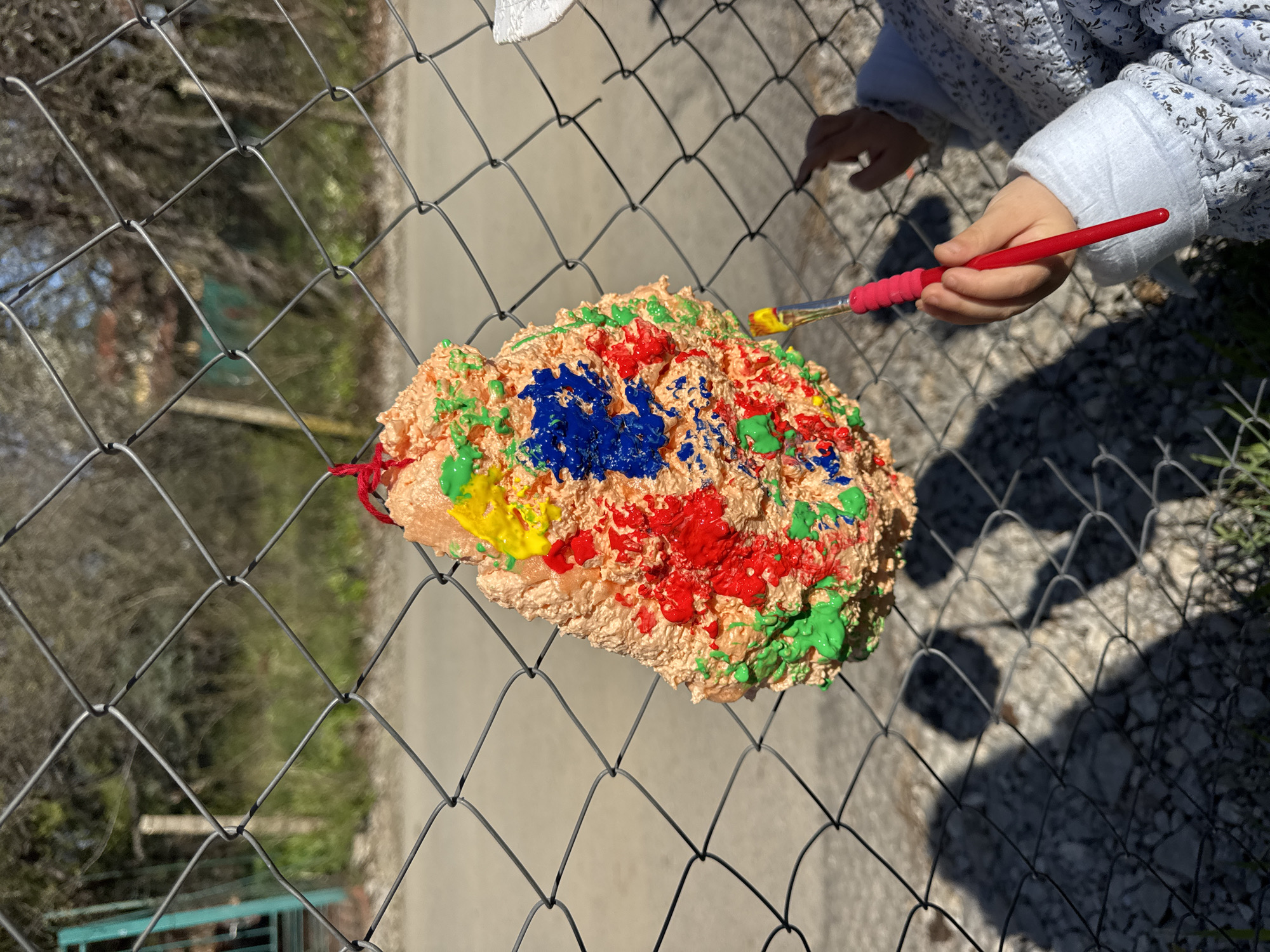A child, wearing a light blue patterned quilted jacket, is painting a colorful object. The object is hung on a chain-link fence. The object is painted with blue, red, yellow, and green paint. The child is using a red-handled paintbrush with yellow paint on the brush. The location appears to be outdoors, possibly in a backyard or park, as suggested by the foliage visible through the fence and the sunny lighting. While it is impossible to definitively confirm the location is in Sofia, Bulgaria, based solely on this image, there is no evidence to indicate otherwise.