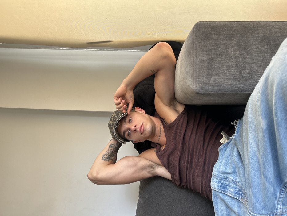 A man is casually reclined on a gray couch, wearing a leopard-print cap, a brown ribbed tank top, and light-wash denim jeans. His left arm, adorned with a floral tattoo, is bent behind his head. His right hand adjusts the cap, drawing attention to his facial features: light eyes and a hint of stubble. A tattoo is visible on his chest. The image is taken at a rotated angle, emphasizing the relaxed pose. The background features a plain wall, creating a minimalist setting.