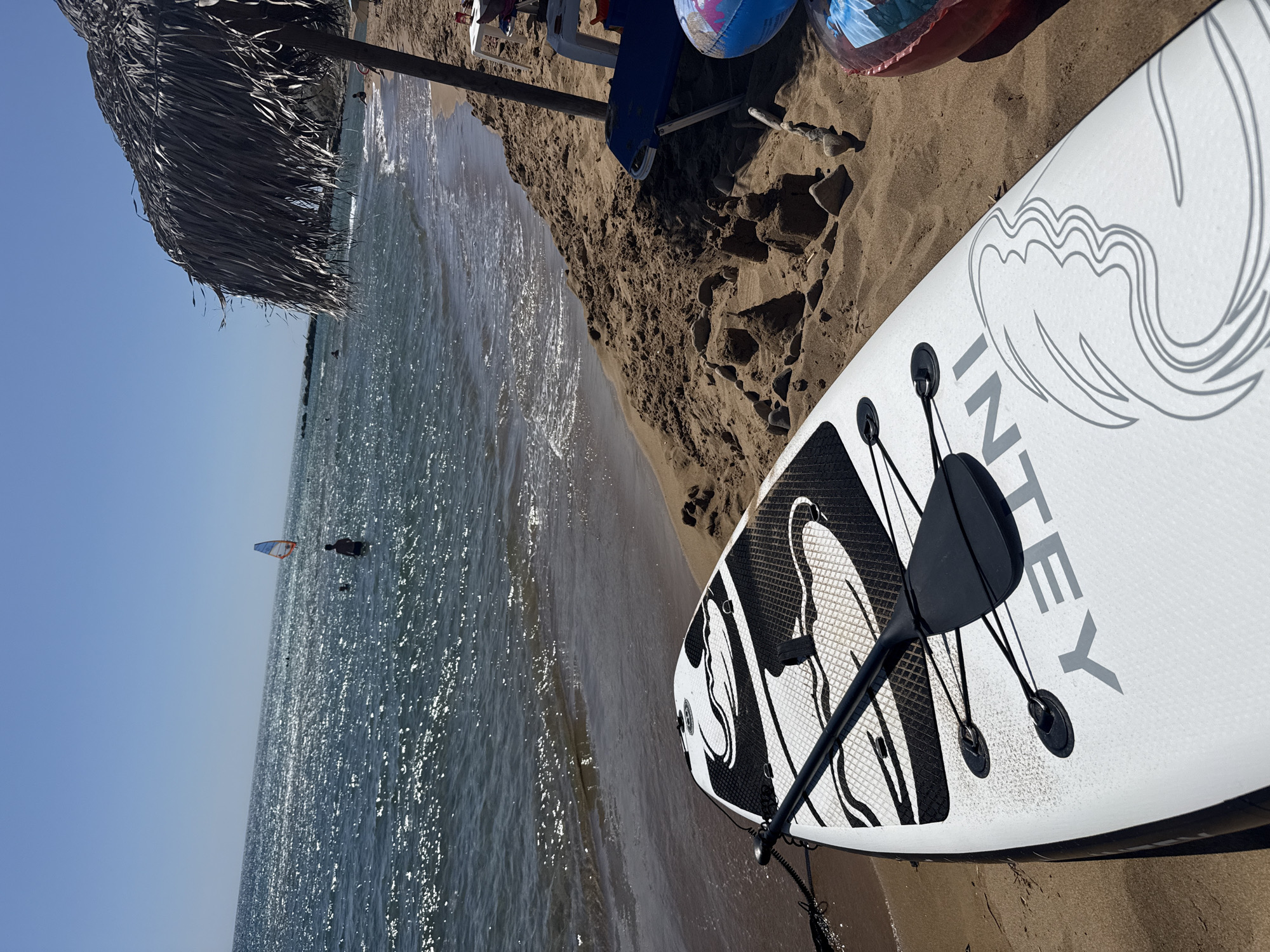 A bright, sunny day at a beach in Municipal Unit of Lefkimmi, Greece, is depicted. In the foreground, a white paddleboard with black trim and a black paddle resting on it is positioned on the sandy shore. The paddleboard has a graphic design with the word "INTEY" visible in grey letters. To the right of the paddleboard, there are sandcastles, stones, a blue lounge chair, a small white table with a red object on it, and part of an inflatable ring. A large thatched umbrella provides shade. In the background, the calm, sparkling sea stretches to the horizon under a clear blue sky. Several people are visible in the water: one person is wading near the shore, and further out, another person is windsurfing with a colorful sail. The gentle waves lap the sandy beach.